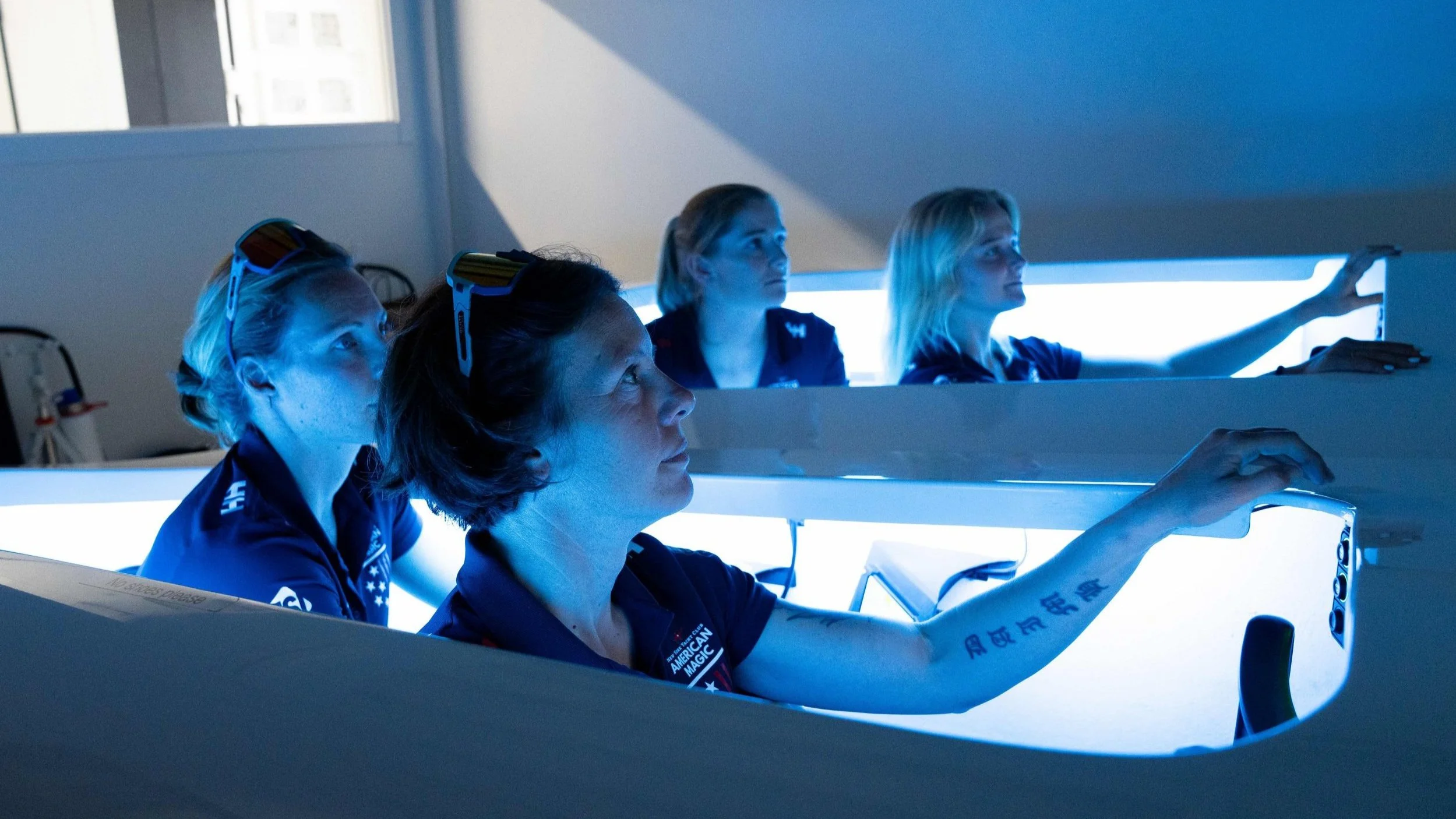 Four women astronauts in blue jumpsuits with NASA logos and American flags, participating in a training session in a mock-up spacecraft, illuminated by blue light.