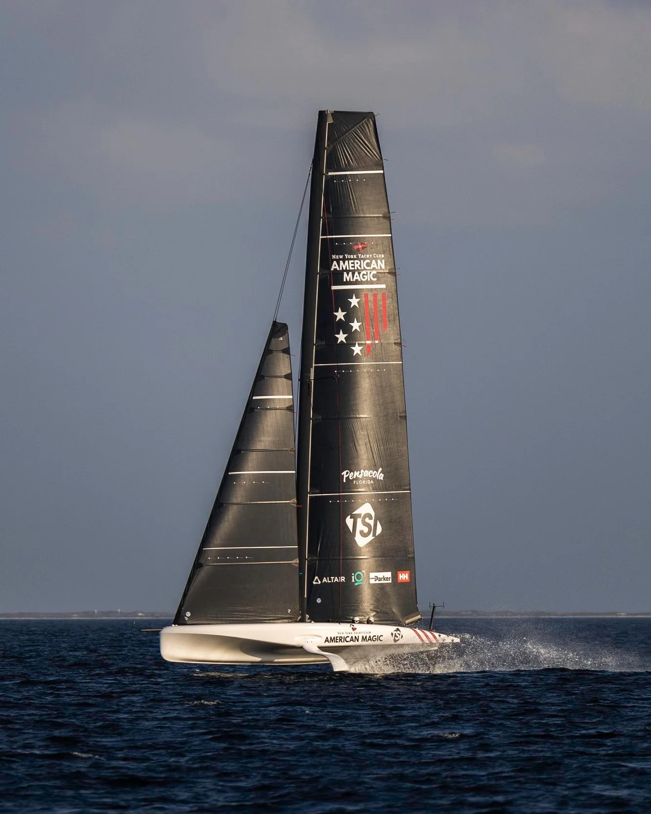 A black sailing yacht named American Magic racing on the water with a dark sky background.