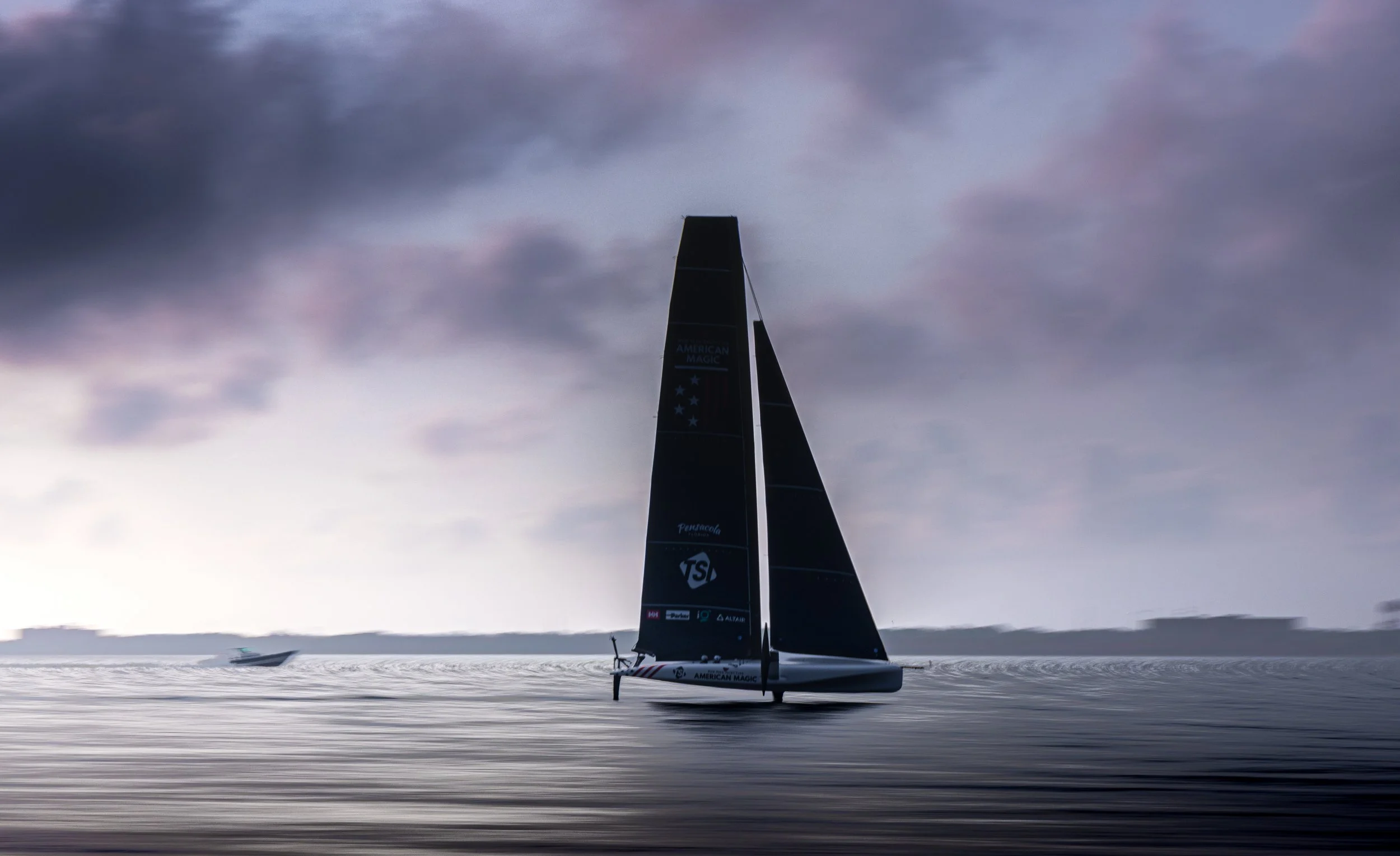 A black sailboat on the water with dark clouds in the sky, next to a boat in the background, during dusk or dawn.