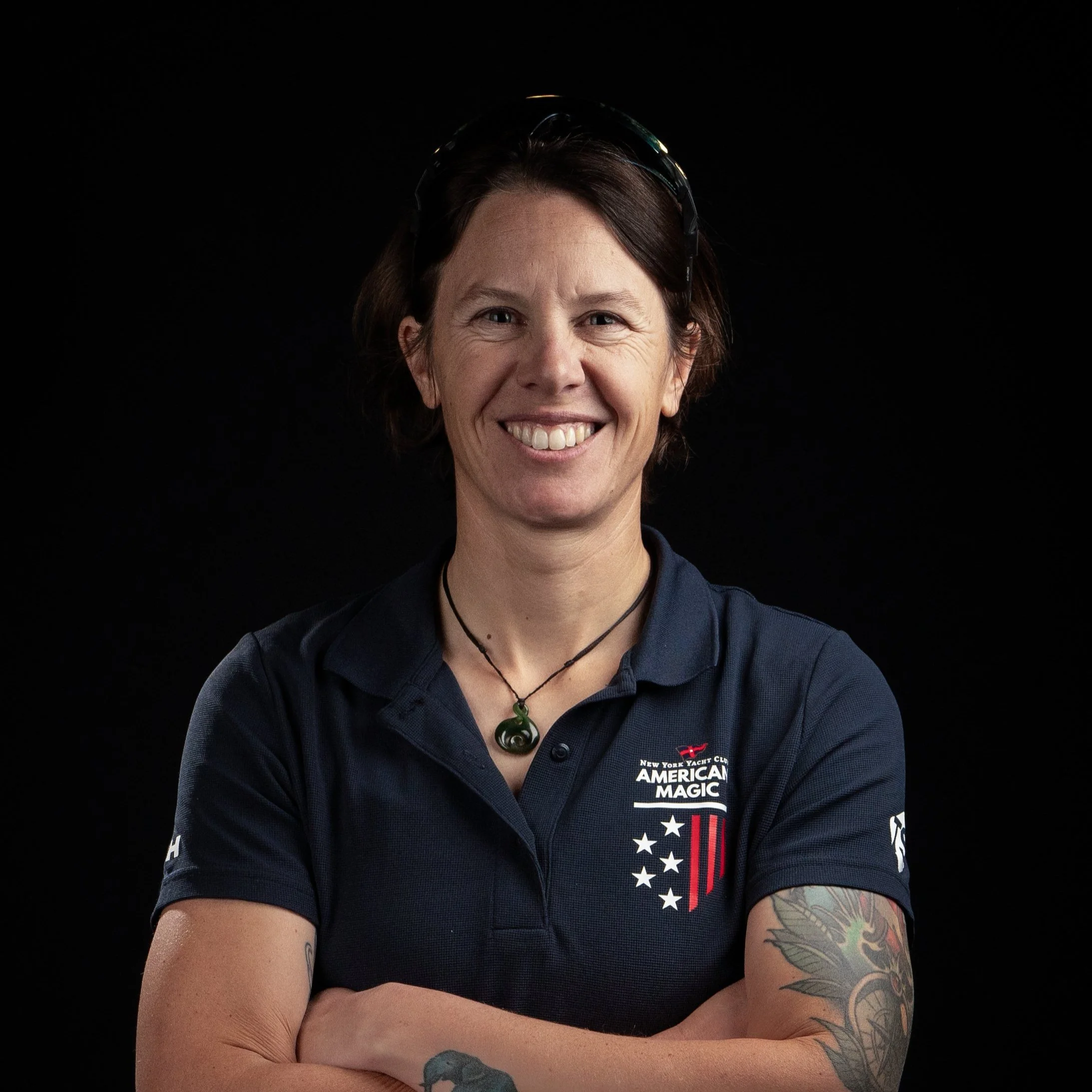 A smiling woman with short dark hair, wearing a dark blue polo shirt with American flag and yacht club logos, tattoos on her arms, and a necklace with a green pendant, against a black background.