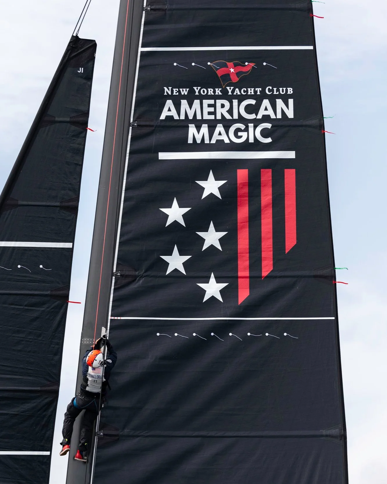 A sailor climbs a black mast at the New York Yacht Club American Magic sailing team event, with the team's logo, stars, and red stripes on the sail.