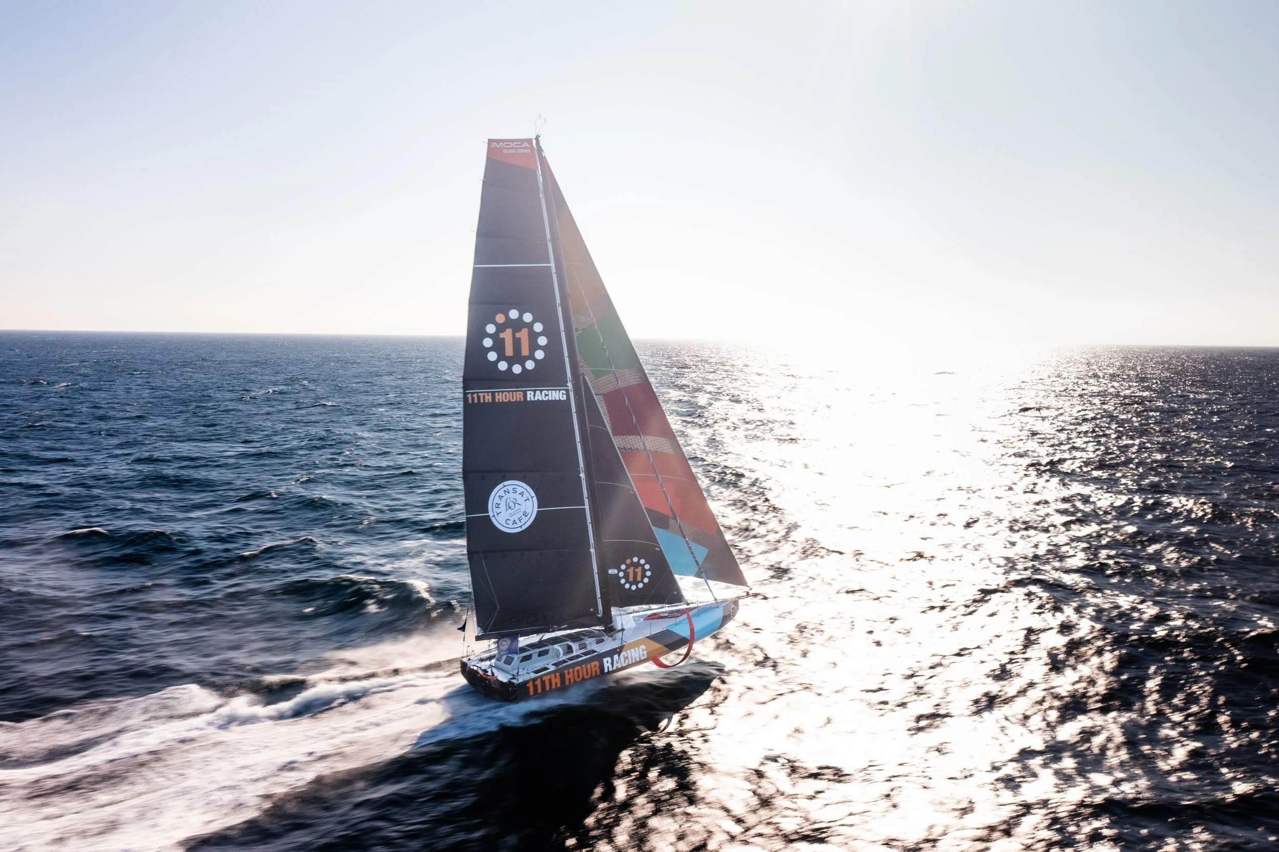 A racing sailboat with black sails on the open ocean during sunset, labeled '11th Hour Racing' and 'Transat Jacques Vabre'.