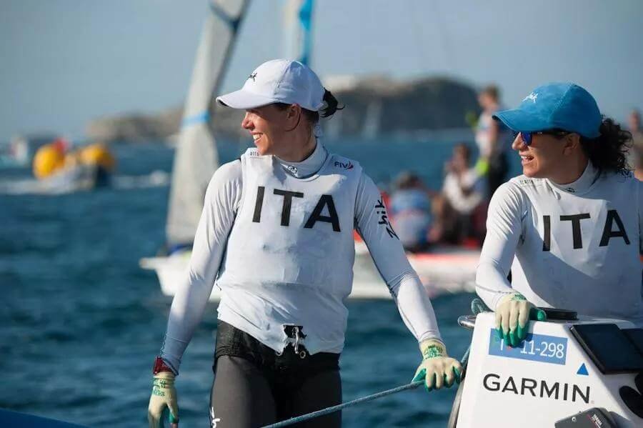 Two women in white sailing jerseys with 'ITA' on them, wearing caps and gloves, smiling on a boat during a sailing race with other boats and yachts in the background.