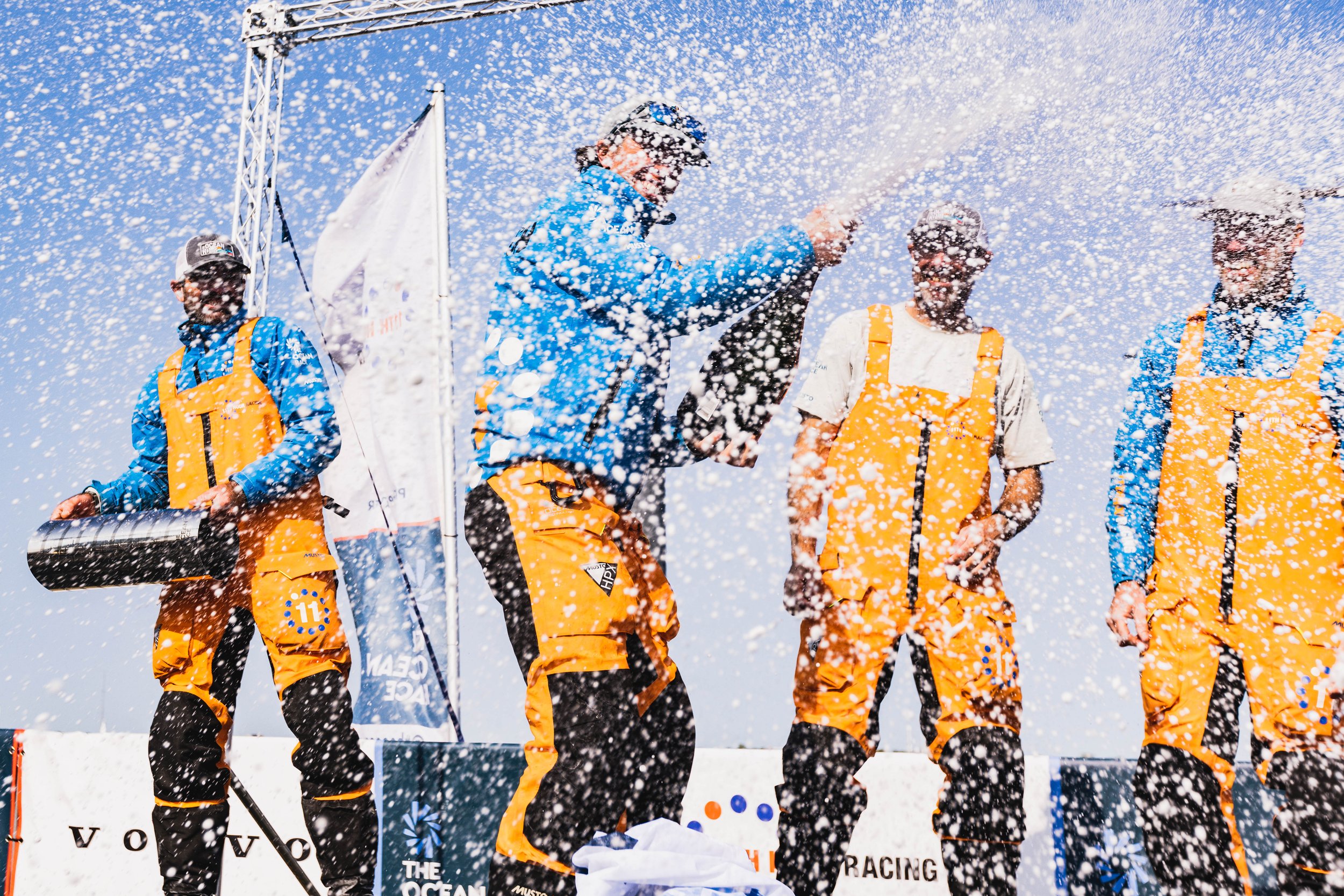 A group of people on a podium celebrating after a race, spraying champagne and wearing orange and blue racing gear.