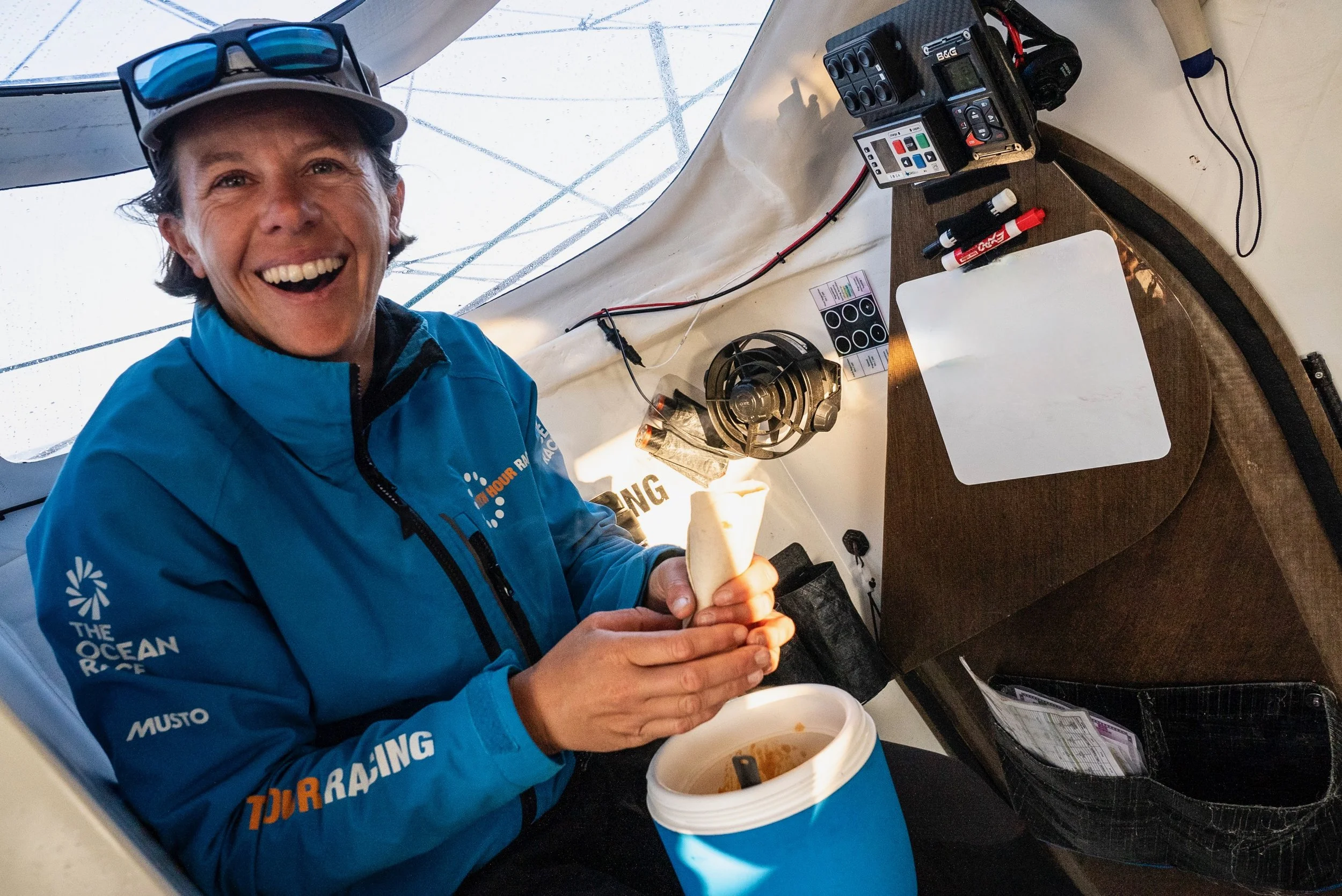 Francesca, with a broad smile, sits at the look out onboard Mālama in The Ocean Race 2022-23 with a blue pot of food on her lap