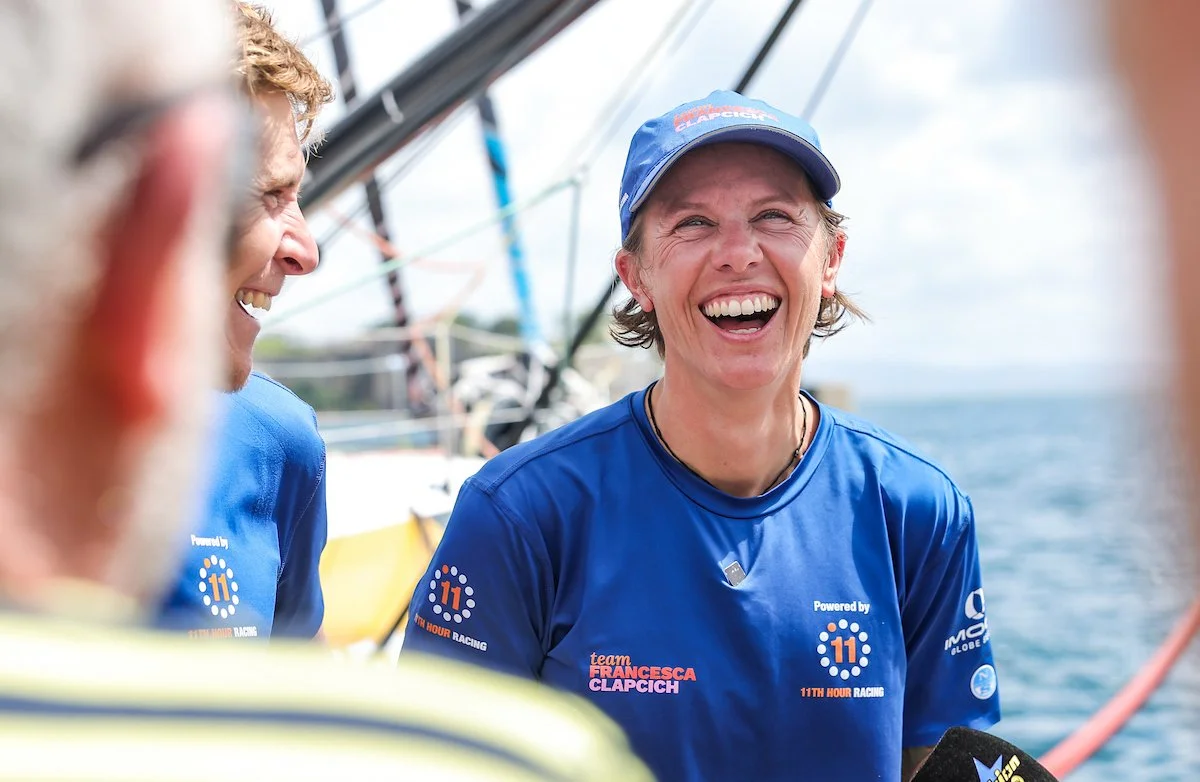 Due donne con magliette blu della squadra e un berretto, che sorridono e ridono su una barca durante una regata.
