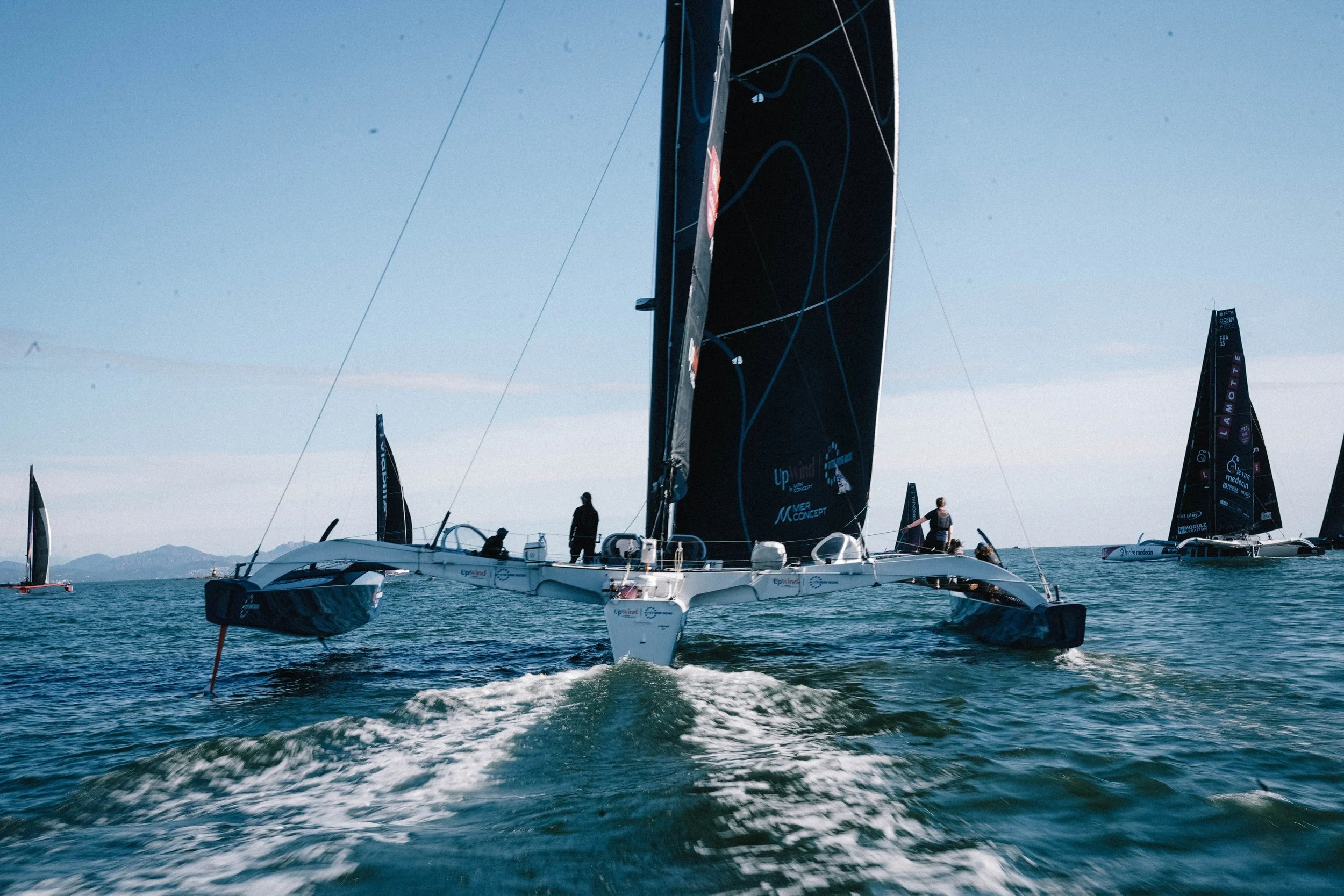 A large racing sailboat with black sails and crew members on board, sailing in open water with other sailboats in the background on a sunny day.