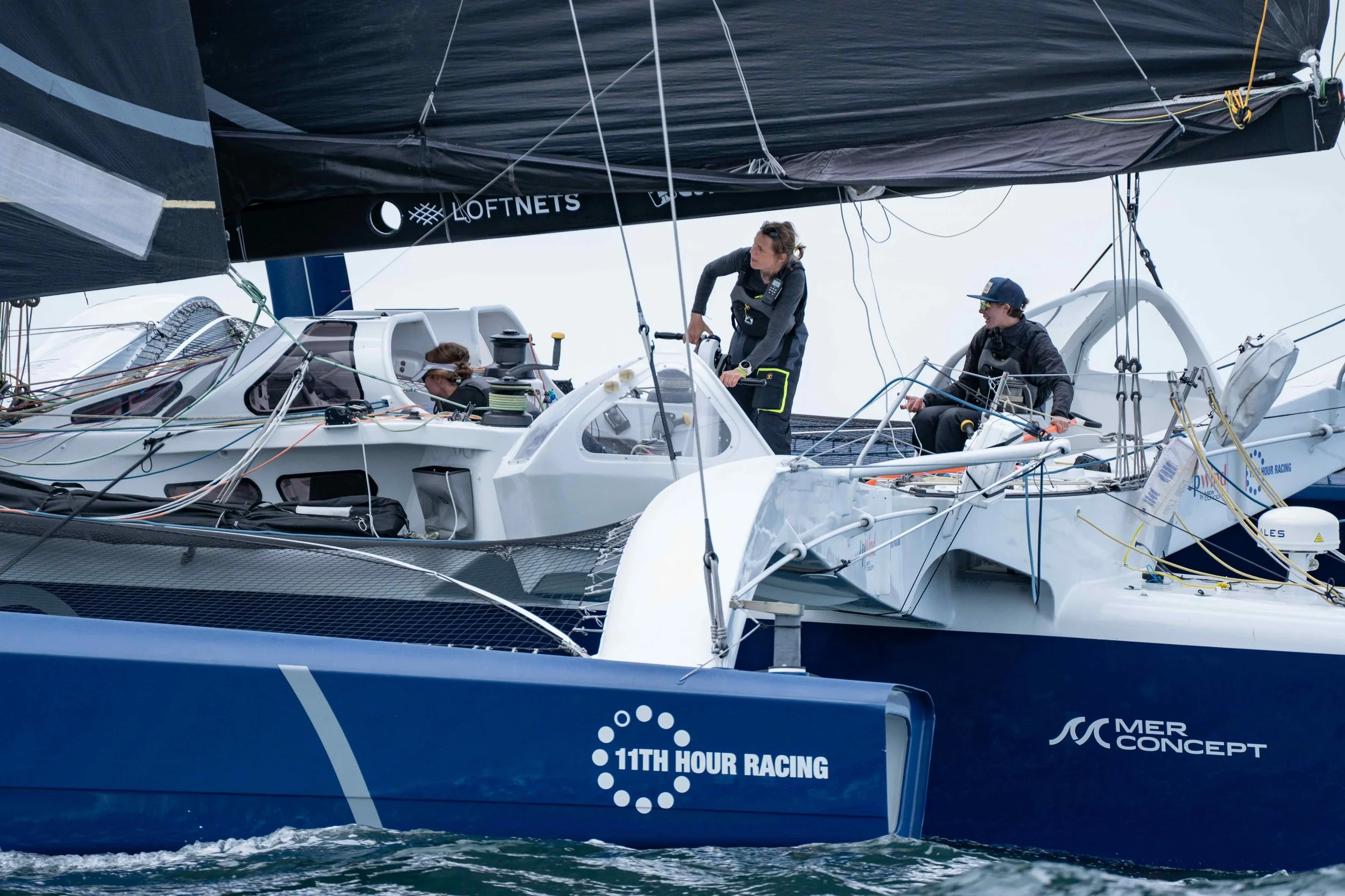 A sailing yacht in a race with crew members on deck, and the boat labeled '11th Hour Racing' and 'Mer Concept'.