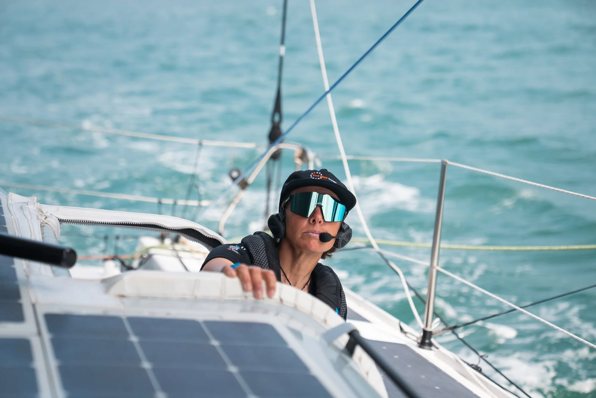Woman sailing on a boat with solar panels, wearing reflective sunglasses, a black cap, and a headset with microphone, on a body of water.