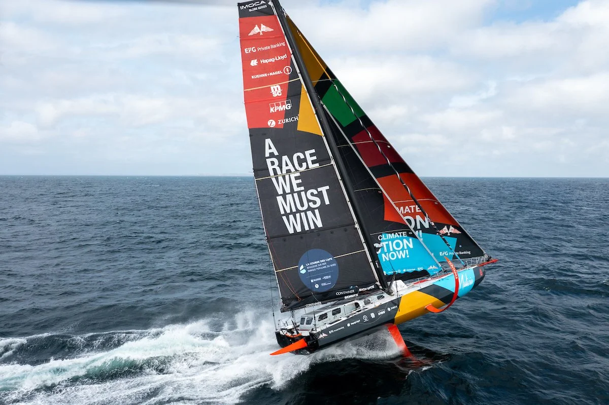 A racing sailboat with black, yellow, green, red, and blue sails on the ocean, featuring sponsorship logos and the message "A RACE WE MUST WIN."