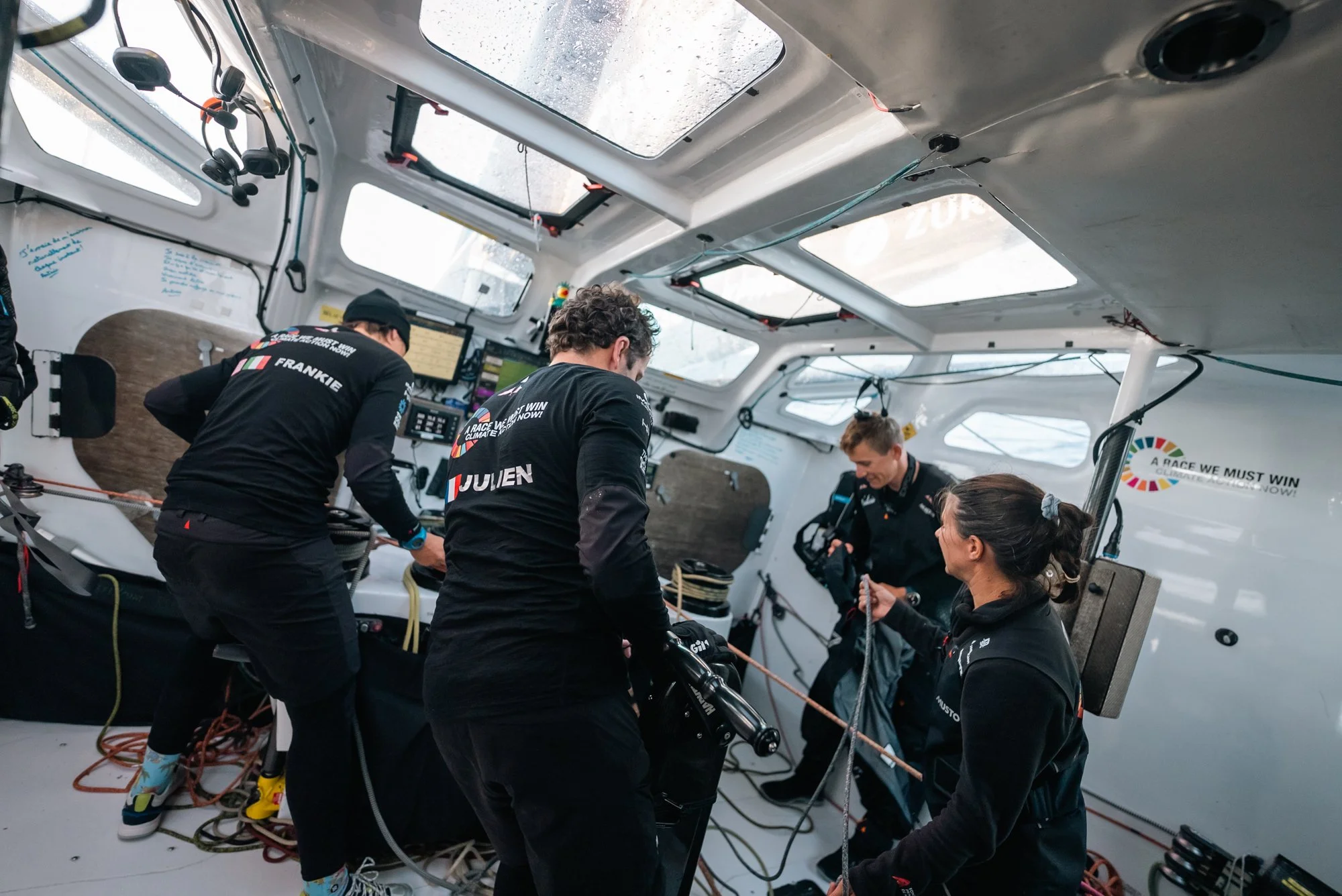 A team of four sailors inside a sailboat, preparing for a race. They are wearing black jackets with names and logos, and are working with ropes and equipment on a bright, well-lit boat with multiple windows.