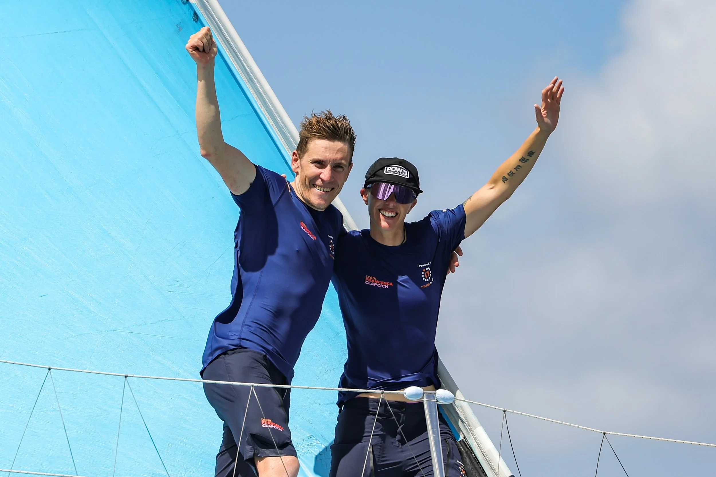 Two smiling sailors in blue shirts celebrating on a sailboat, with one raising his fist and the other extending his arm, against a cloudy sky.