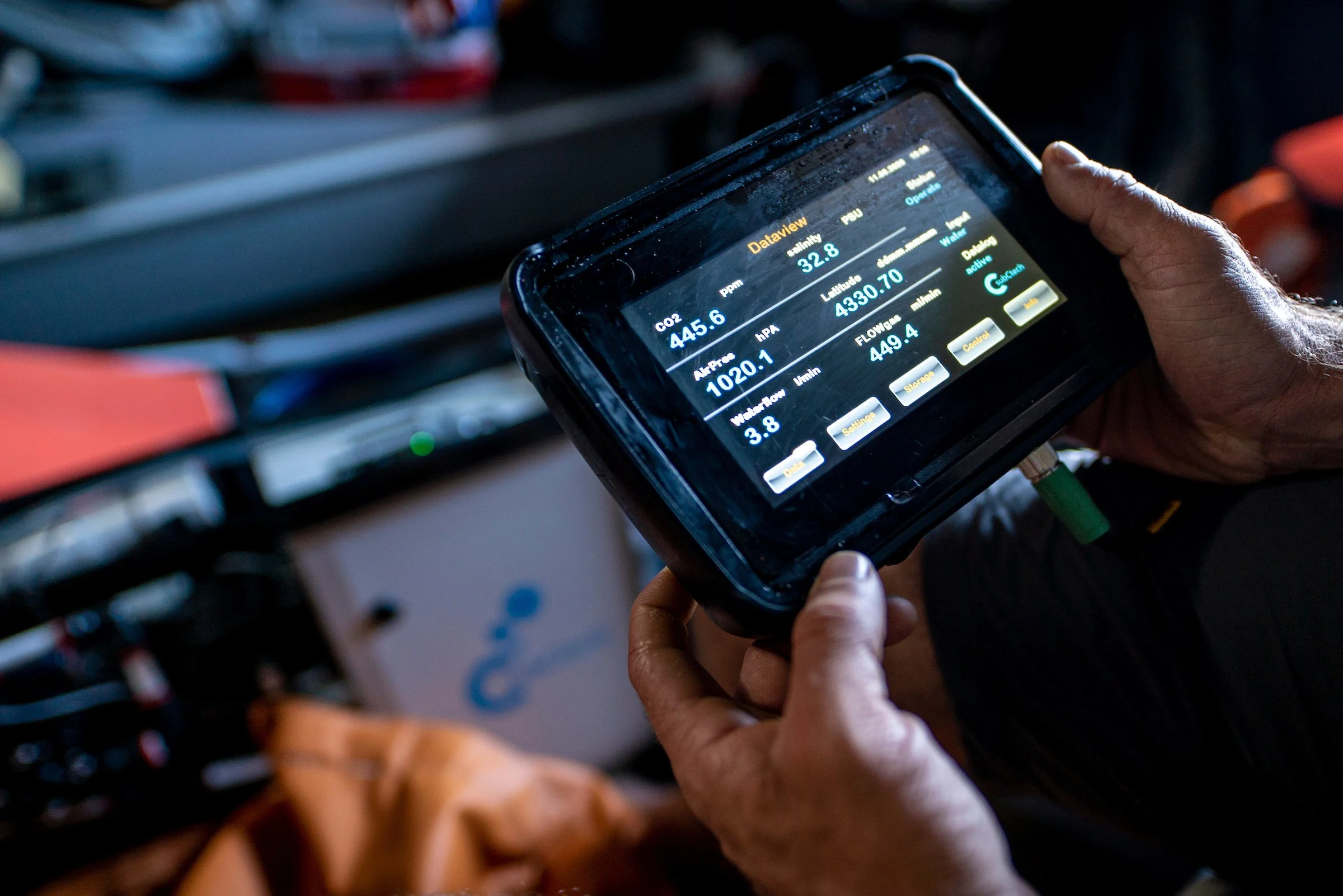 A person holding a black device displaying vital signs such as CO2 level, pH, hydration, and other data, in front of a background with equipment and a blue box.