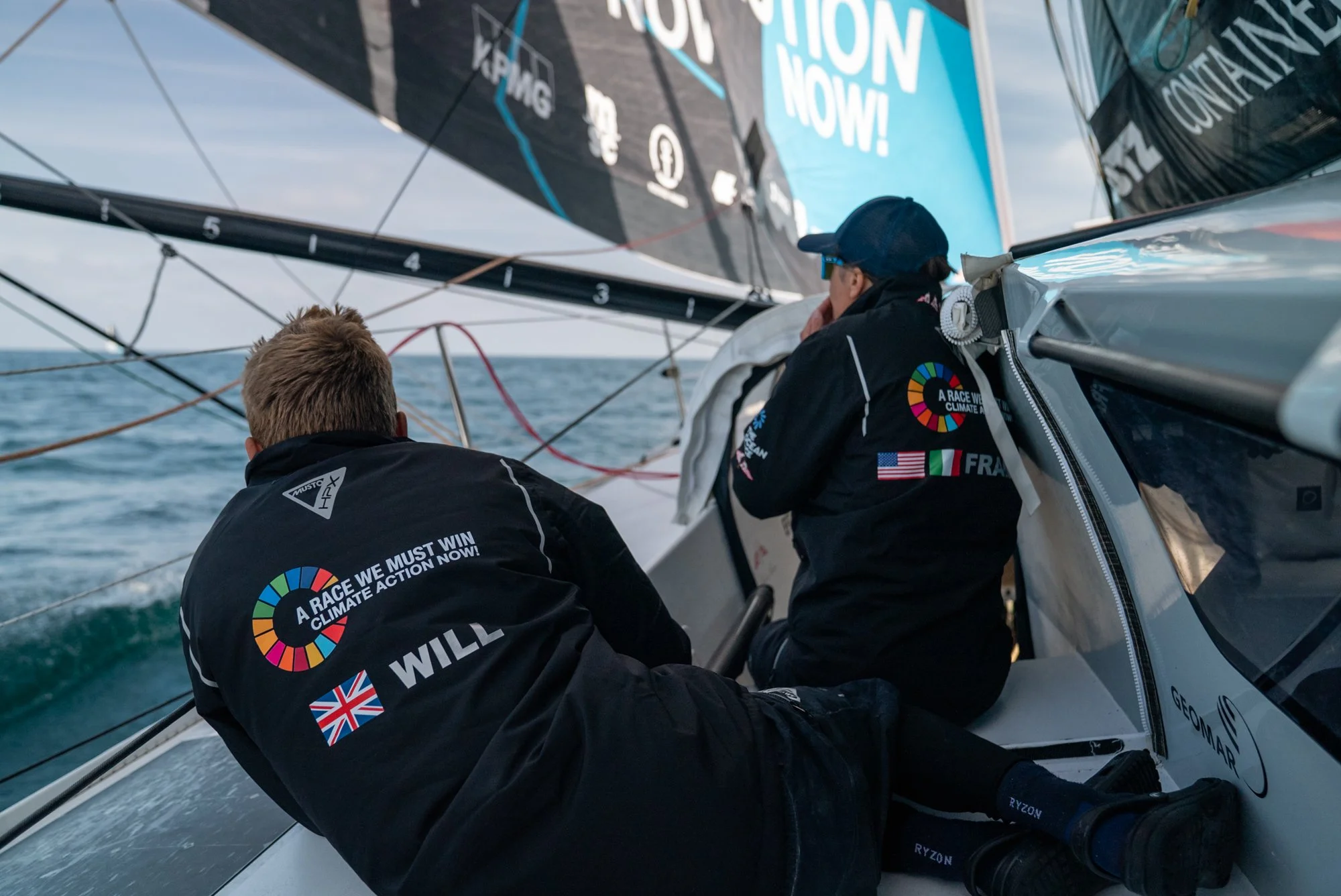 Two sailors on a sailboat during a race, wearing black jackets with climate action slogans, one with a British flag and the other with an American flag, on the open water with the sails in the background.