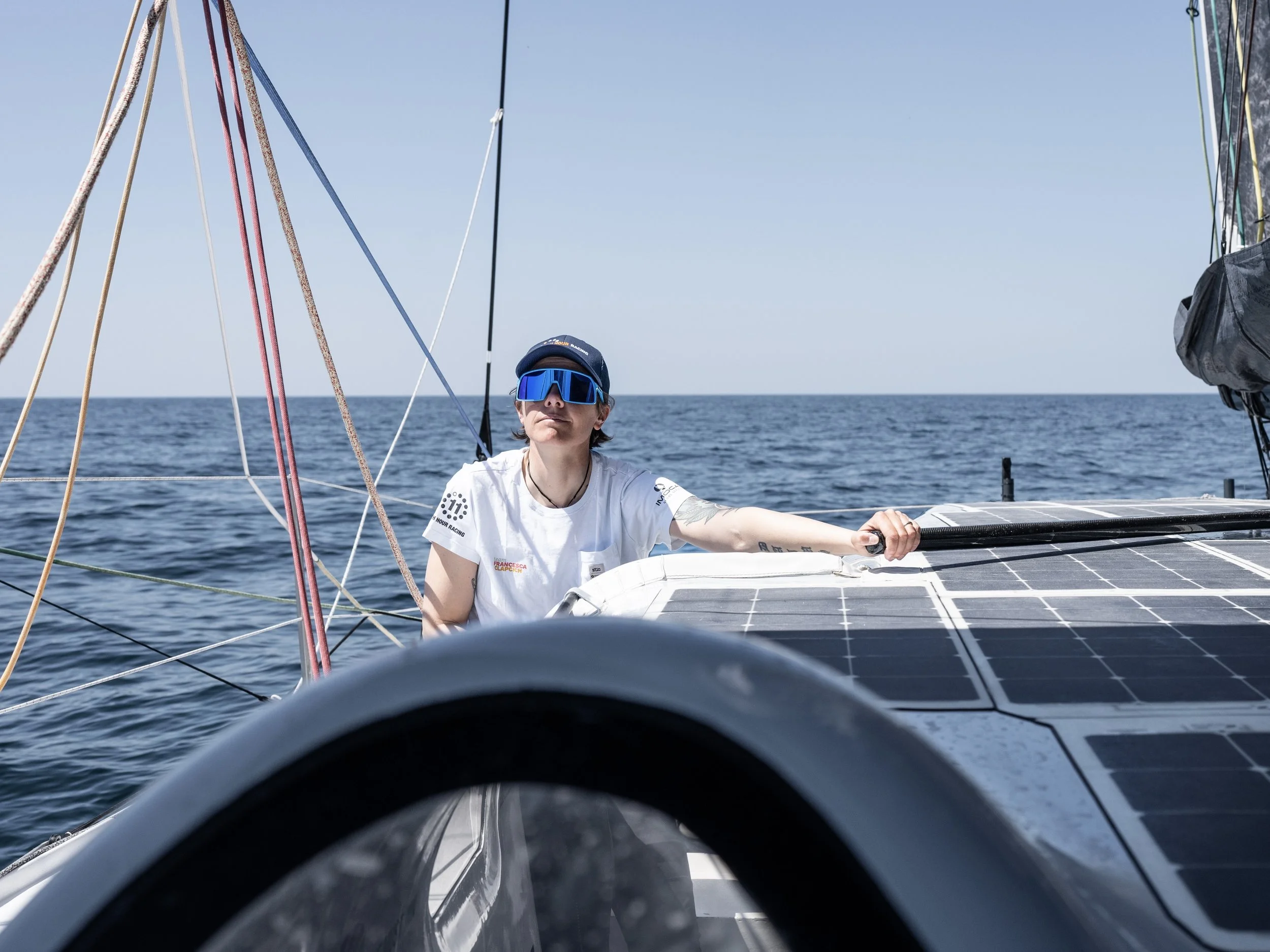 Person wearing sunglasses and a cap on a solar-powered sailboat in open water.