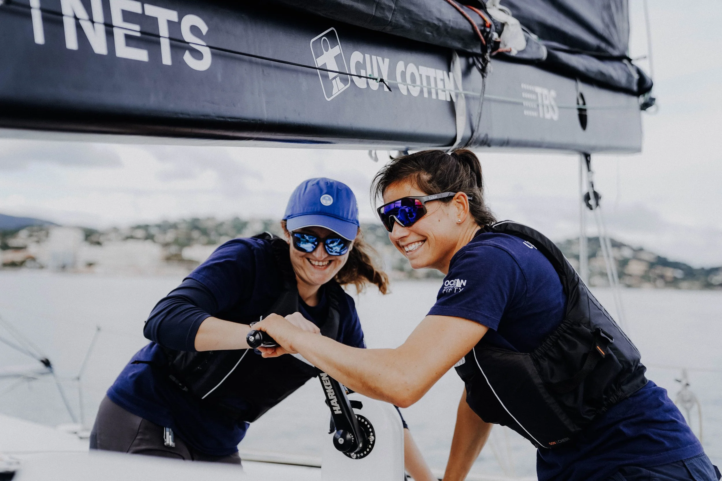 Two women in sunglasses smiling and shaking hands on a sailboat with water and hills in the background.