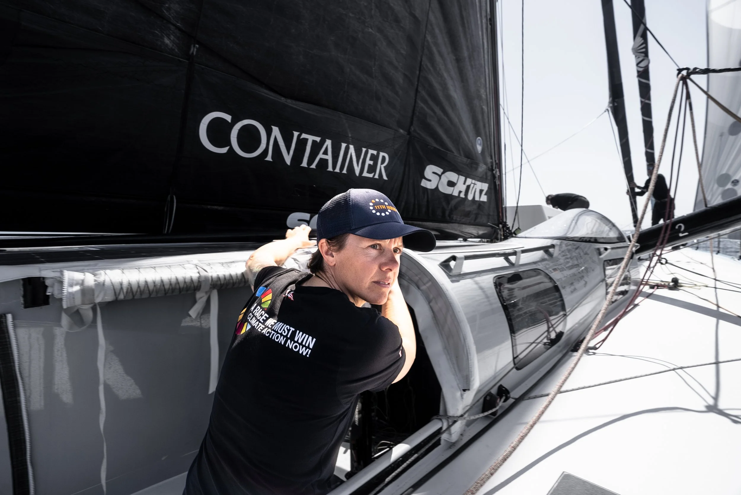A woman working on a sailboat, wearing a navy blue cap and a black t-shirt with climate message, leaning into the boat's interior near the cabin window.
