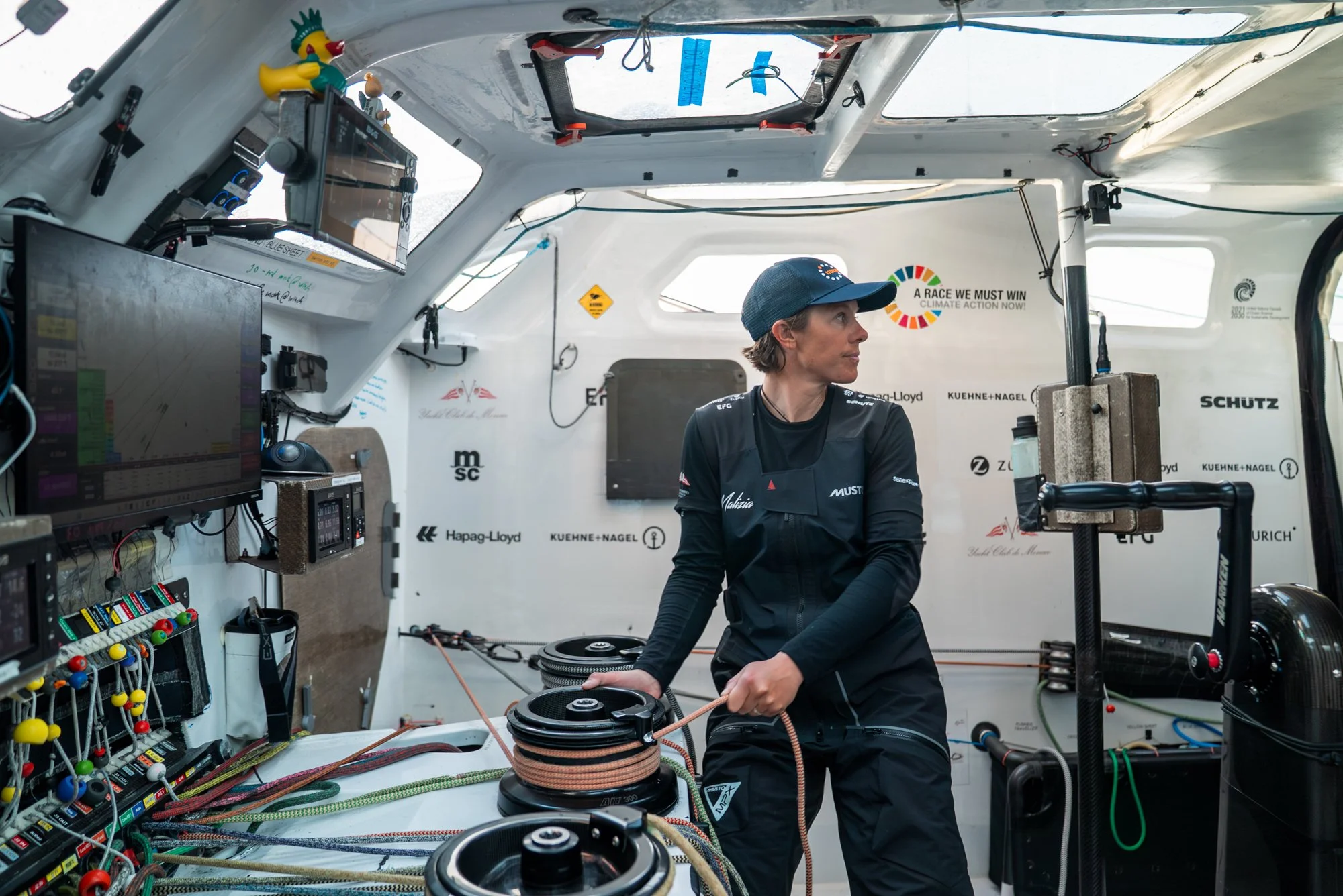 A woman in dark athletic clothing and a cap inside a racing yacht's cabin, surrounded by equipment, ropes, and navigation devices, with climate action slogans on the wall.