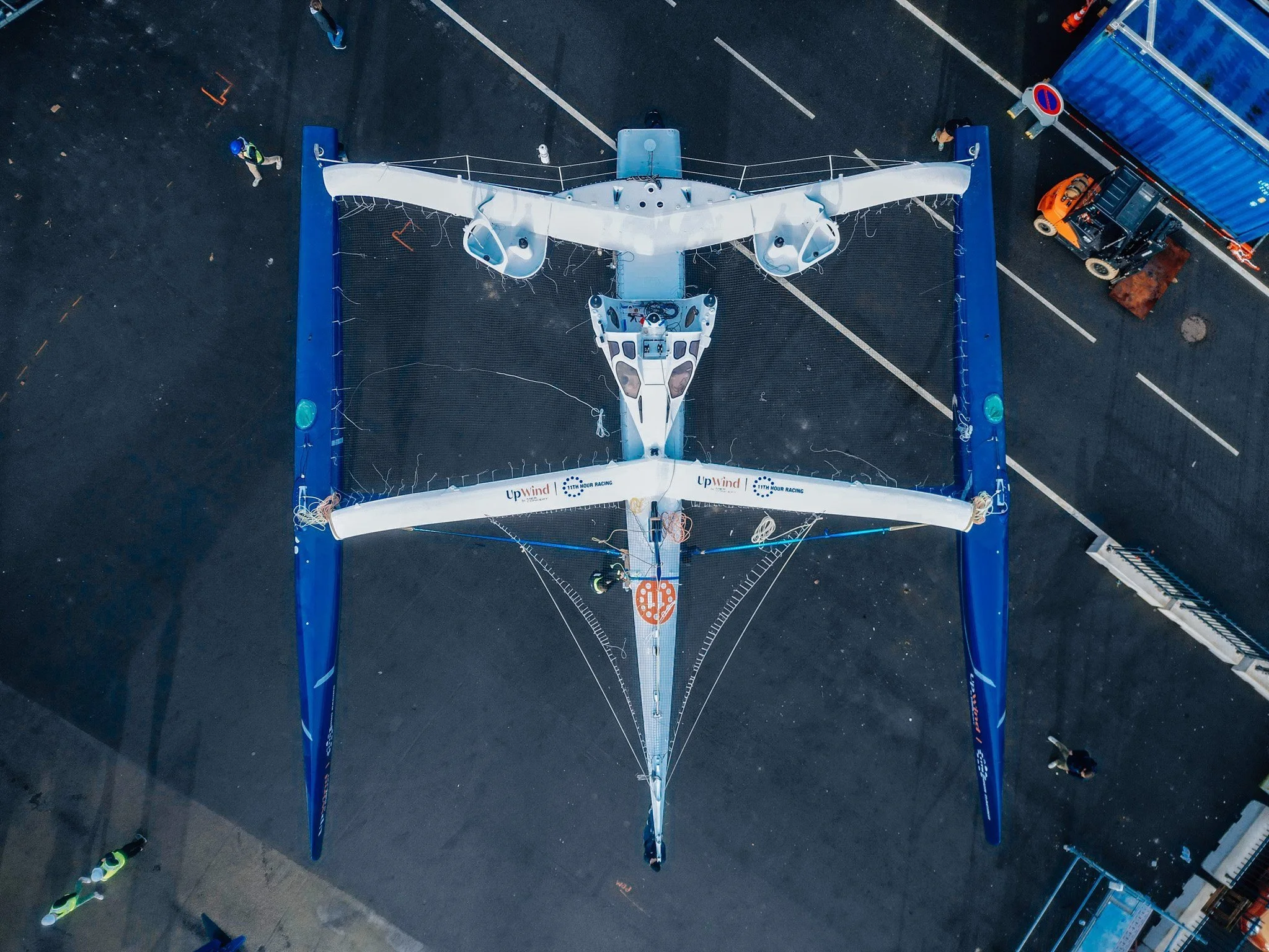 A large sailboat with a modern, aerodynamic design is positioned on land, viewed from above. It has a white and blue color scheme, with the words 'Upwind' and '111th Hour Racing' visible on the hull. The boat is secured with ropes and surrounded by a safety barrier, with personnel nearby preparing for transport or construction.
