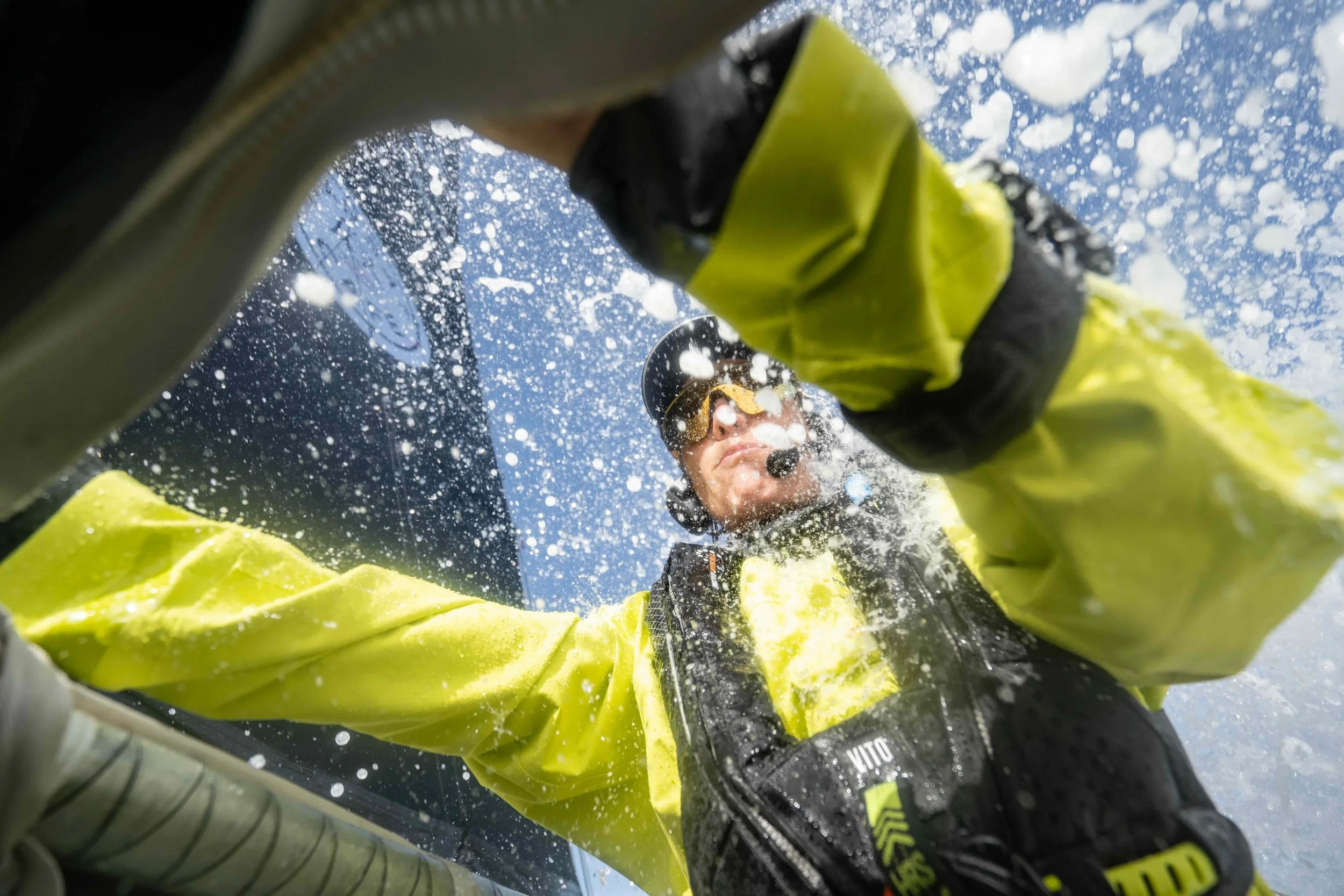 A person in a yellow waterproof jacket and black gloves is cleaning the windshield of a boat with water splashing around, wearing a headset and sunglasses.