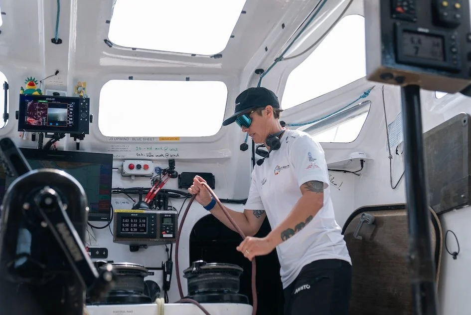 A woman with tattoos, wearing a white shirt, cap, and sunglasses, working with technical equipment inside a space capsule or aircraft.