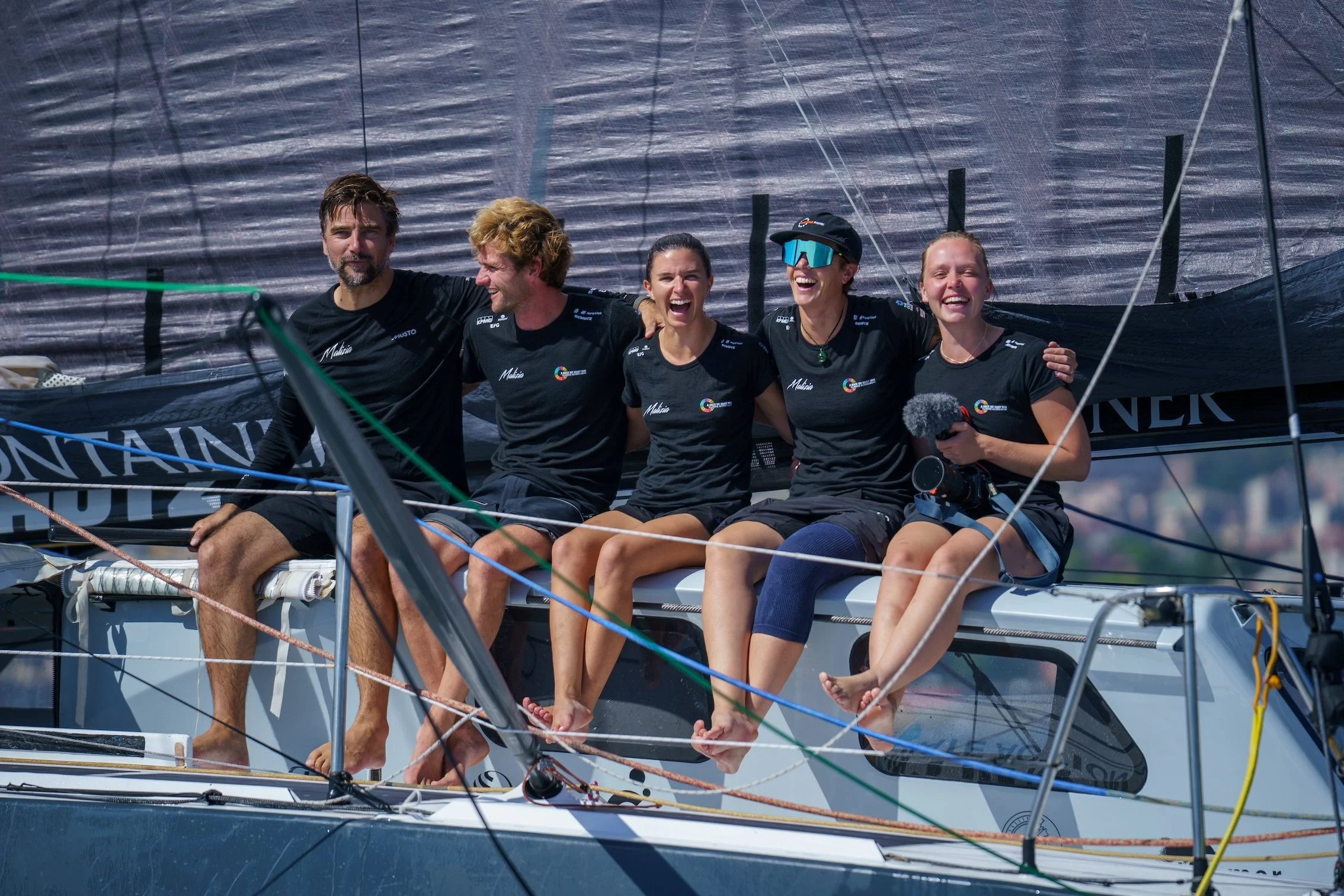 A group of five people sitting on the edge of a sailboat, smiling and laughing, with a large black sail in the background.