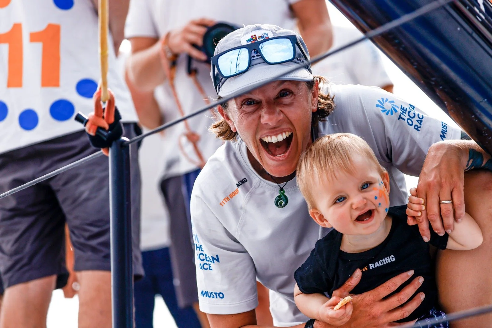 A woman and a young child smiling and leaning close together, with the woman holding the child. The woman is wearing a cap with sunglasses on top, and the child has face paint and is holding a small snack. They are at an outdoor event with other people in the background.