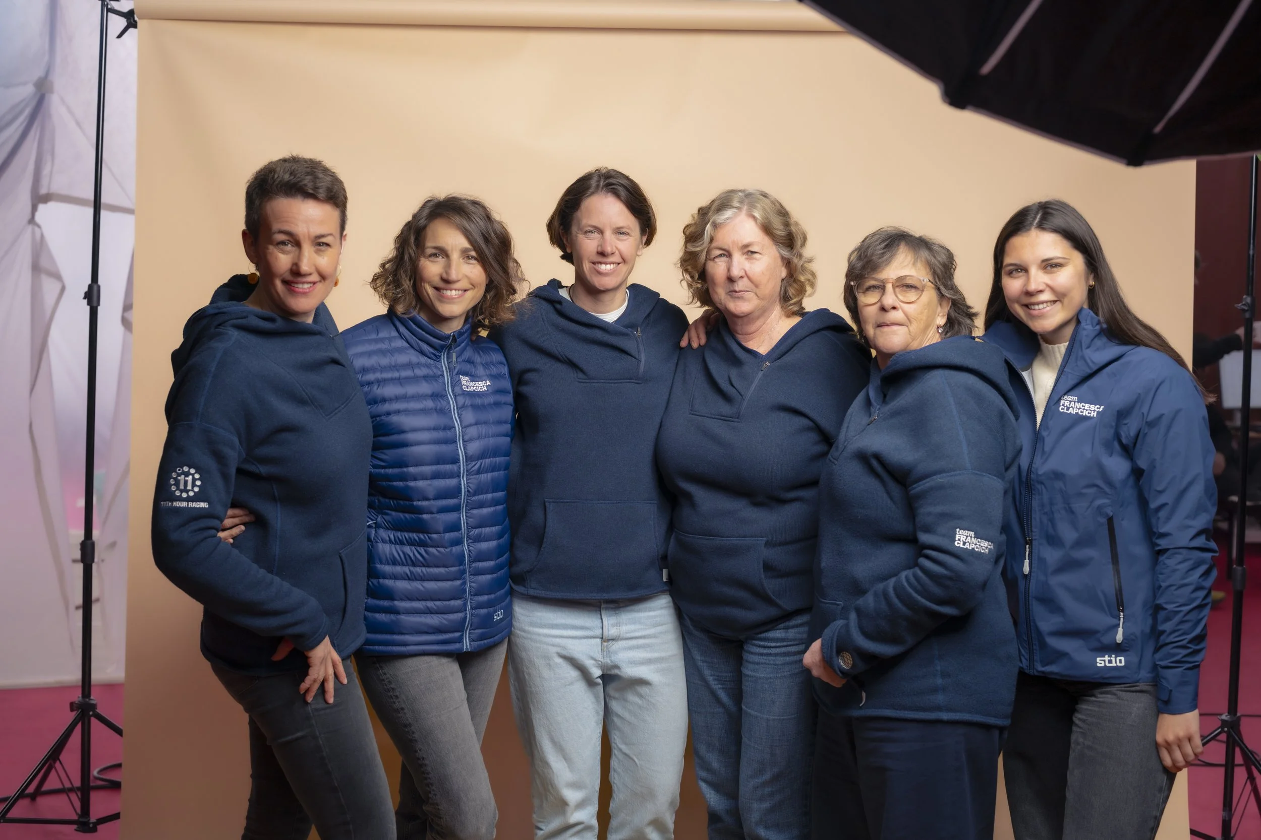 Group of six women standing together in front of a beige background, wearing blue jackets and smiling for the photo.