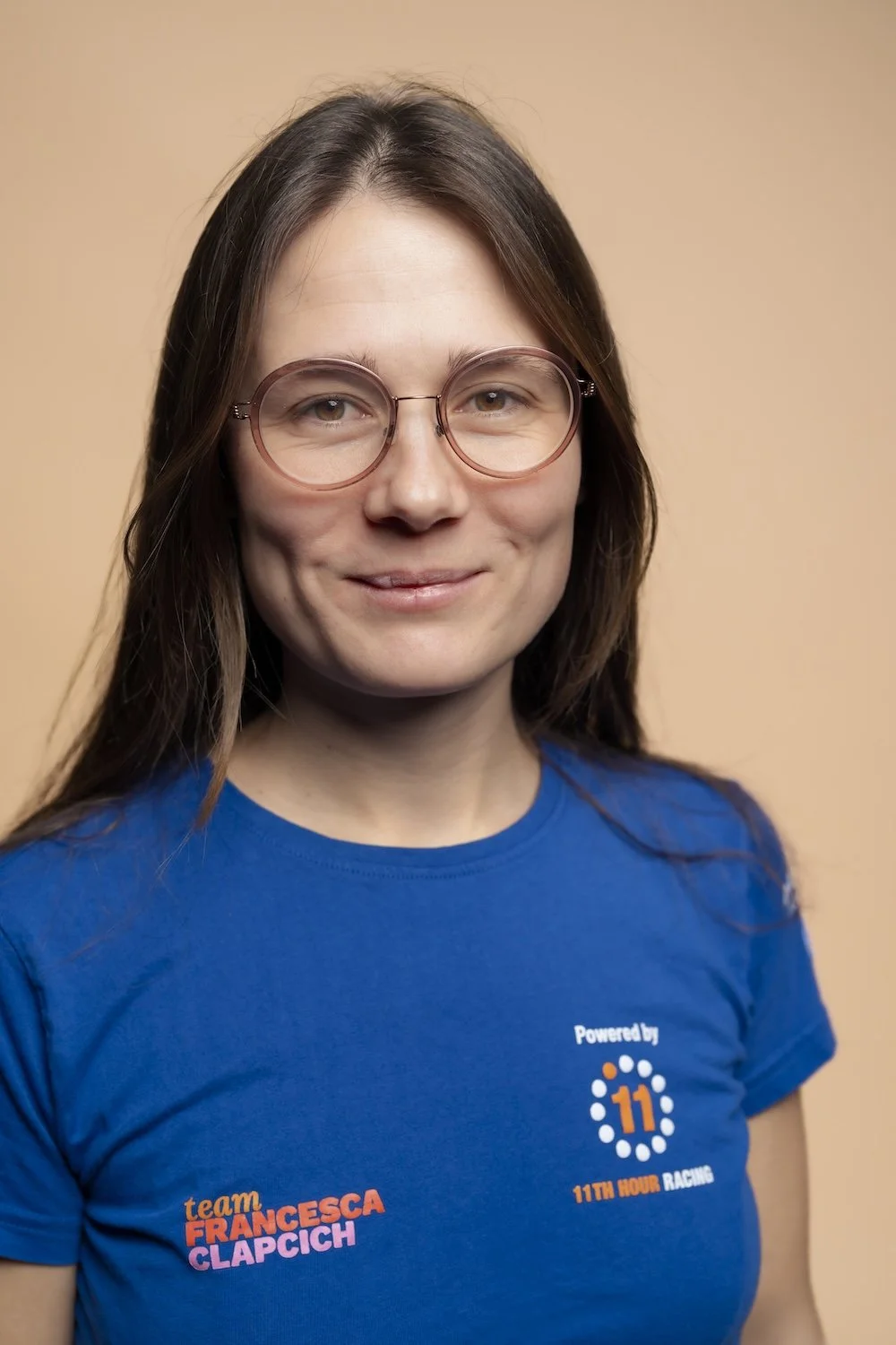 A woman with long brown hair wearing glasses and a blue t-shirt with logos and text, smiling at the camera against a tan background.