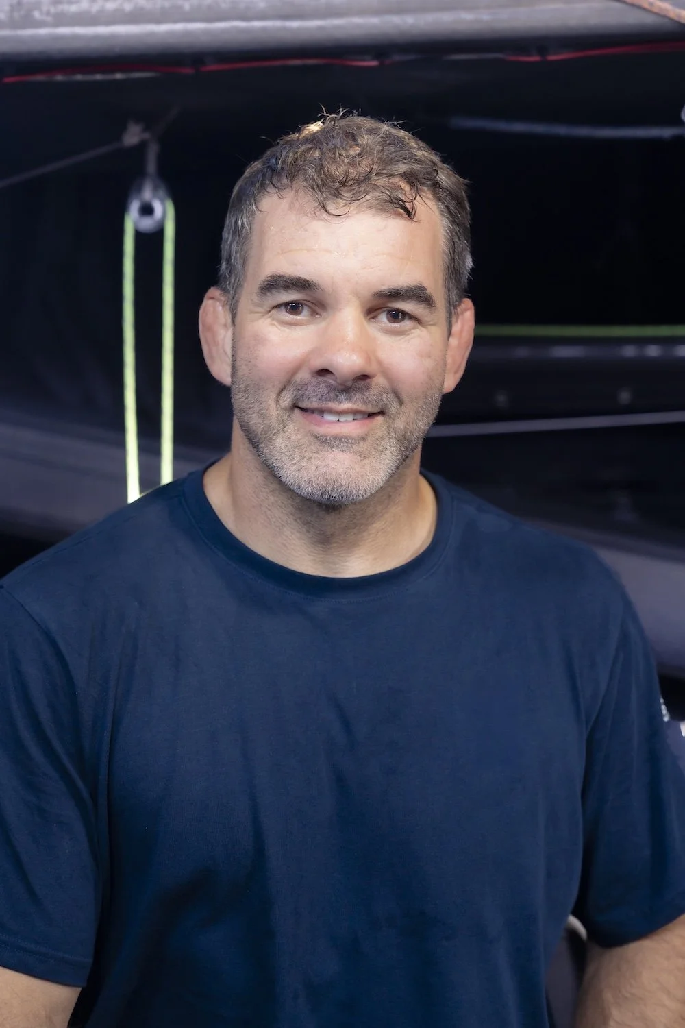 Man with short, curly brown hair and a beard, wearing a navy blue shirt, smiling indoors with a dark background.