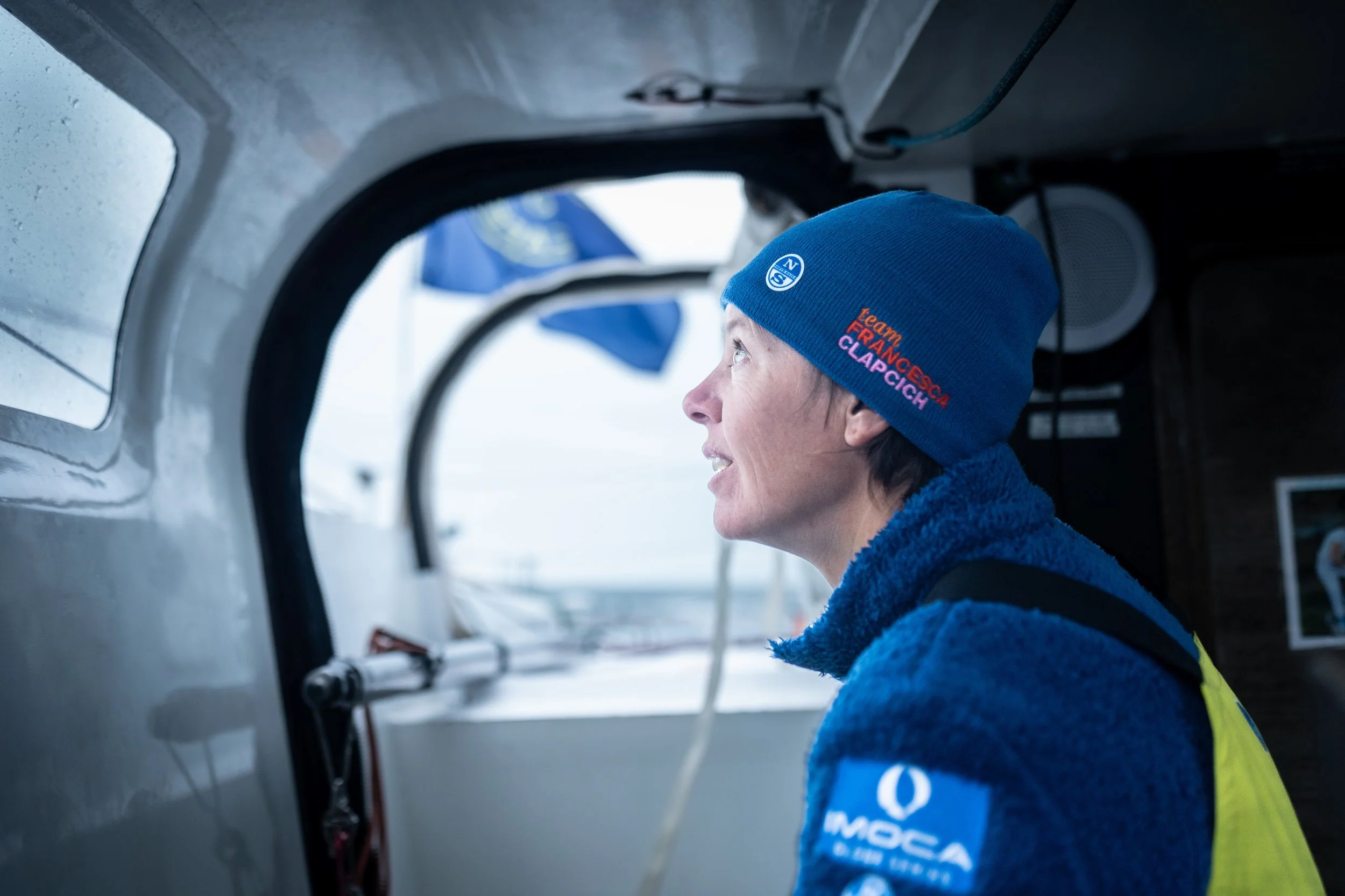 A woman in a blue beanie and blue fleece jacket sitting inside a boat, looking out of a round window with a view of water and a sailboat in the background.