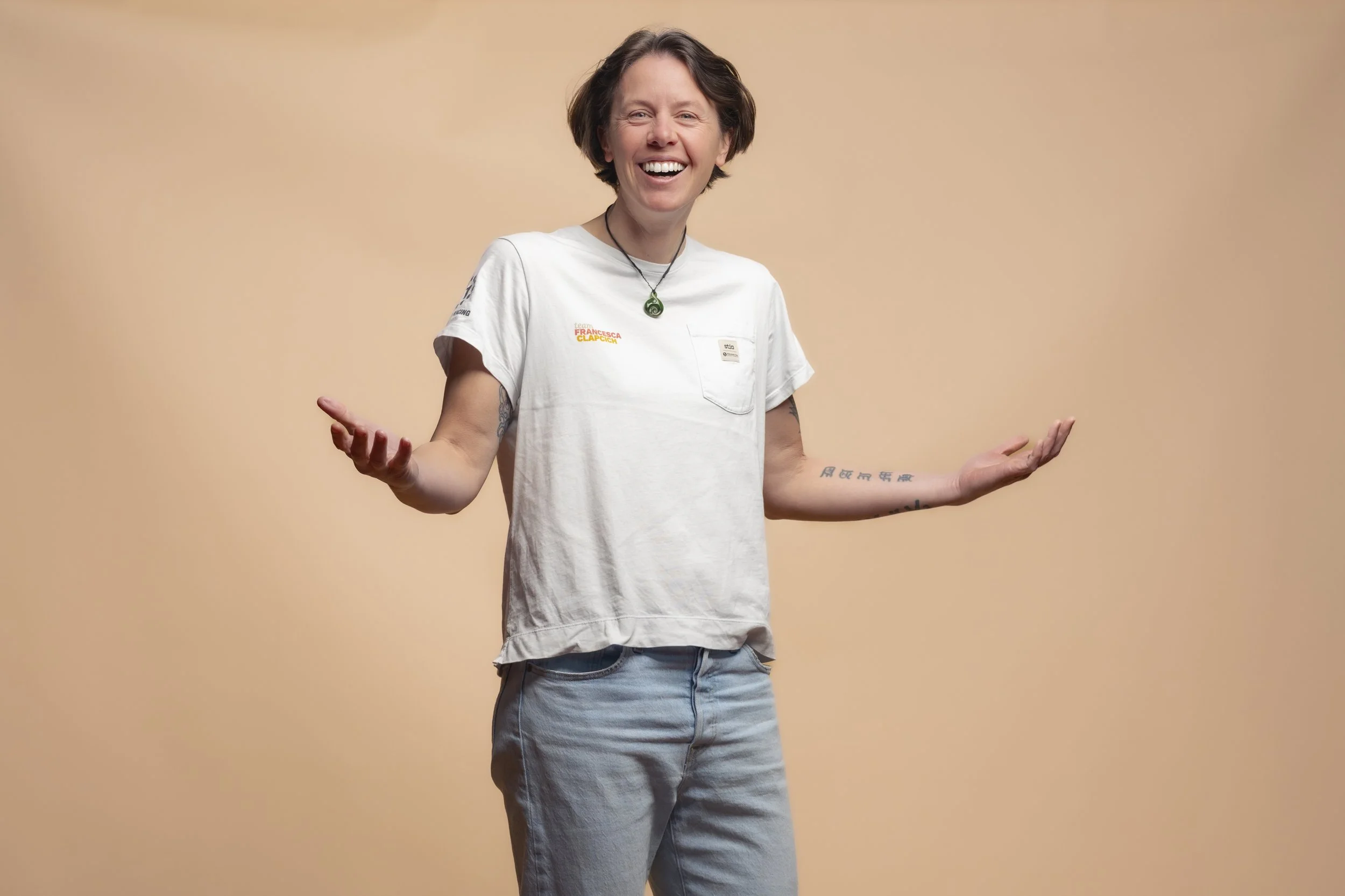 A person with short brown hair and tattoos on their arms, wearing a white T-shirt and light blue jeans, smiling and gesturing with their arms open against a peach-colored background.