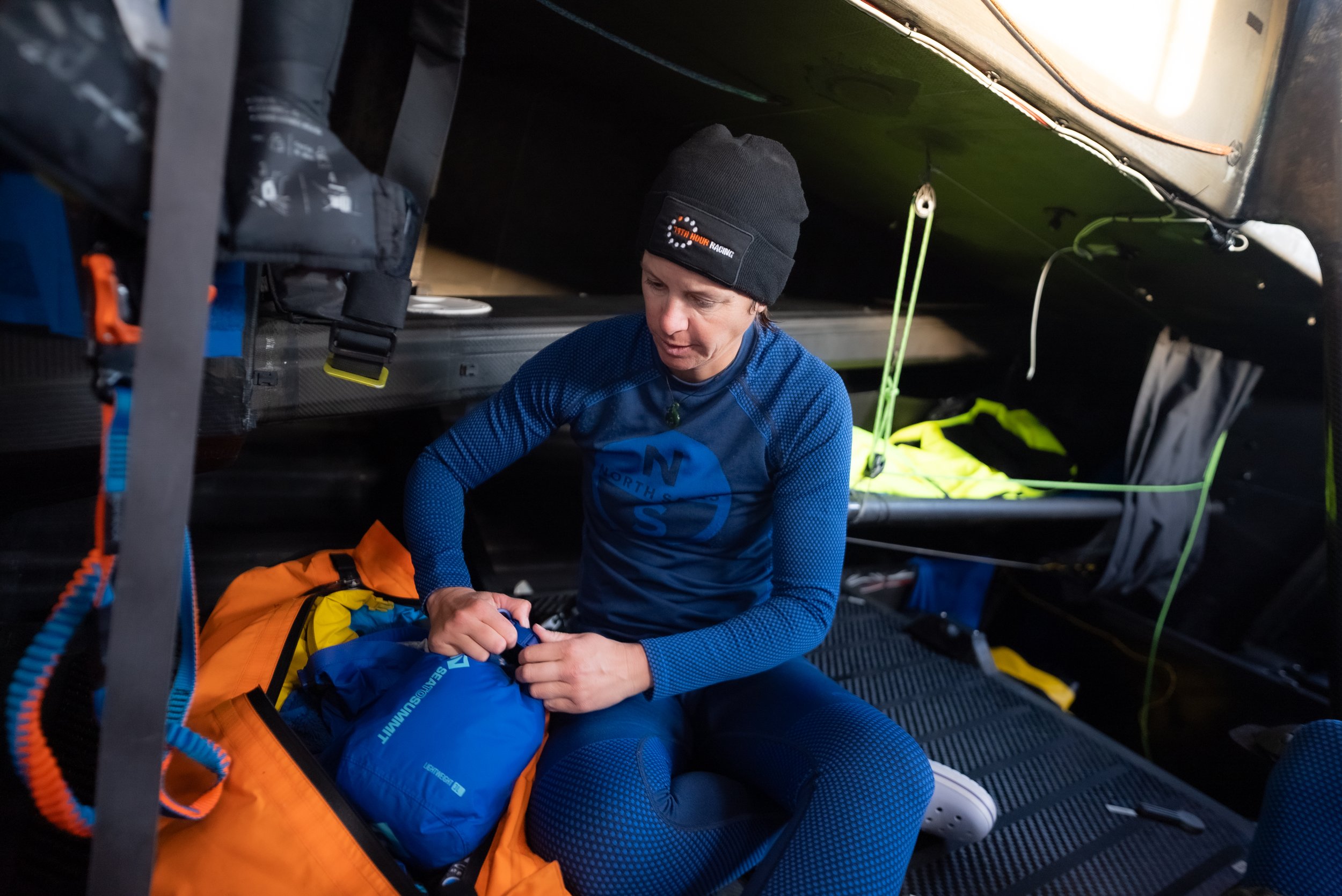 A woman in outdoor gear sitting inside an enclosed vehicle compartment, organizing gear from a backpack. She wears a black beanie and a blue long-sleeve shirt with a nautical compass logo, surrounded by equipment and supplies.