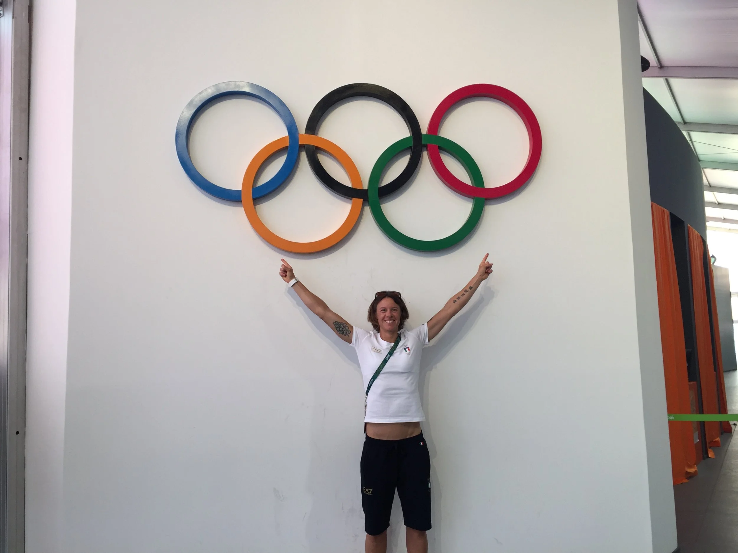 A woman standing with arms raised under the Olympic rings logo at the Olympics venue.