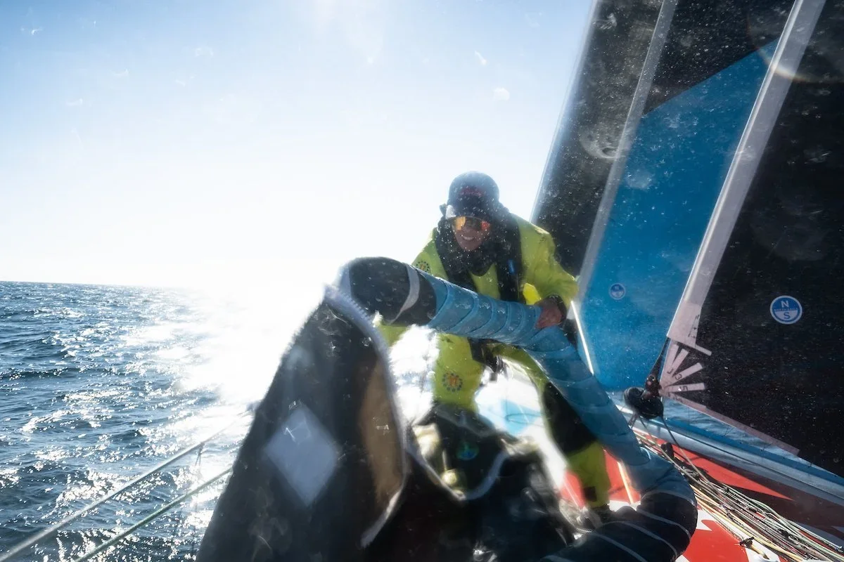 A person in a yellow jacket and black helmet leaning over the side of a sailboat during daytime, with the ocean and bright sky in the background.