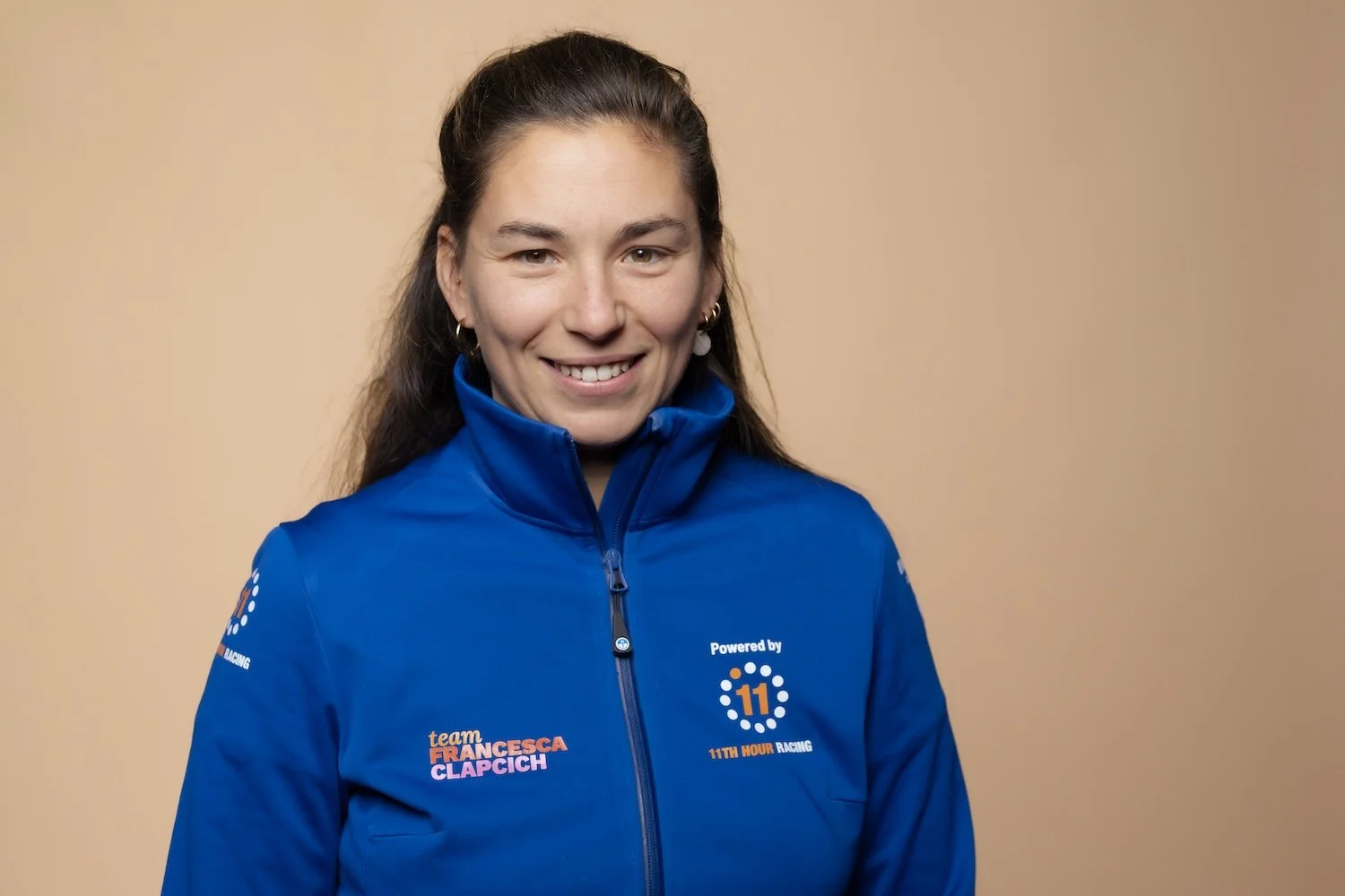 A woman smiling, wearing a blue racing jacket with team logos and text, standing against a plain beige background.
