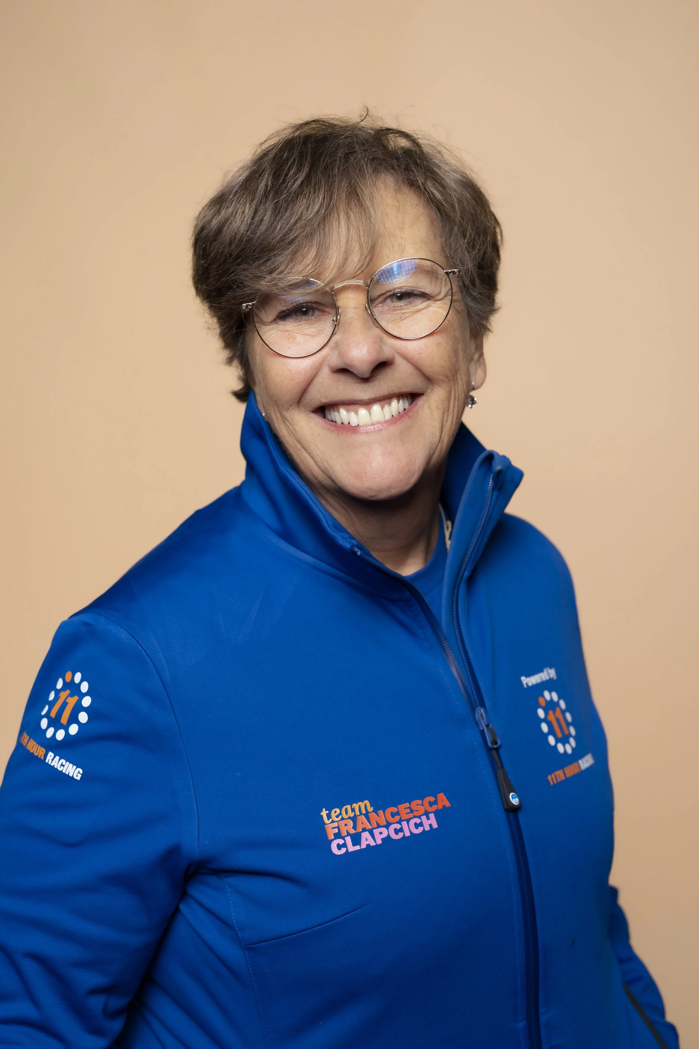 A smiling woman with short brown hair, glasses, and wearing a blue zip-up jacket with the text 'team FRANCESCA CLAPCICH' and logos on the chest and sleeve, standing against a beige background.