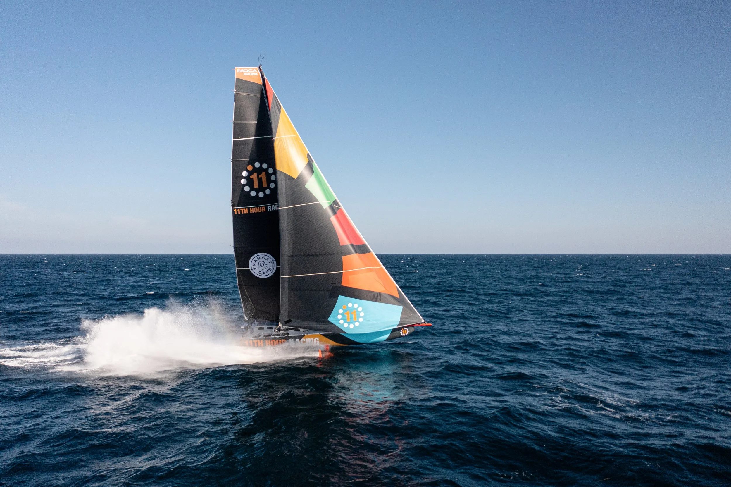 A racing sailboat speeding across the ocean with a colorful sail against a clear blue sky.