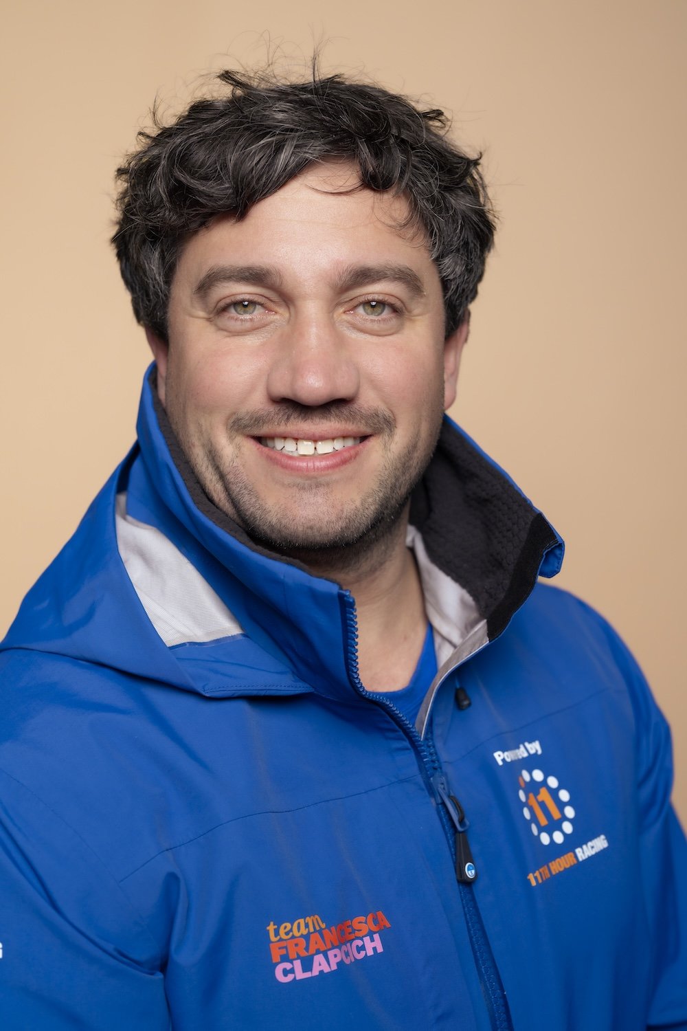 Portrait of a smiling man with dark, curly hair wearing a blue jacket with logos for 11th Hour Racing and team Francesca Clappich.