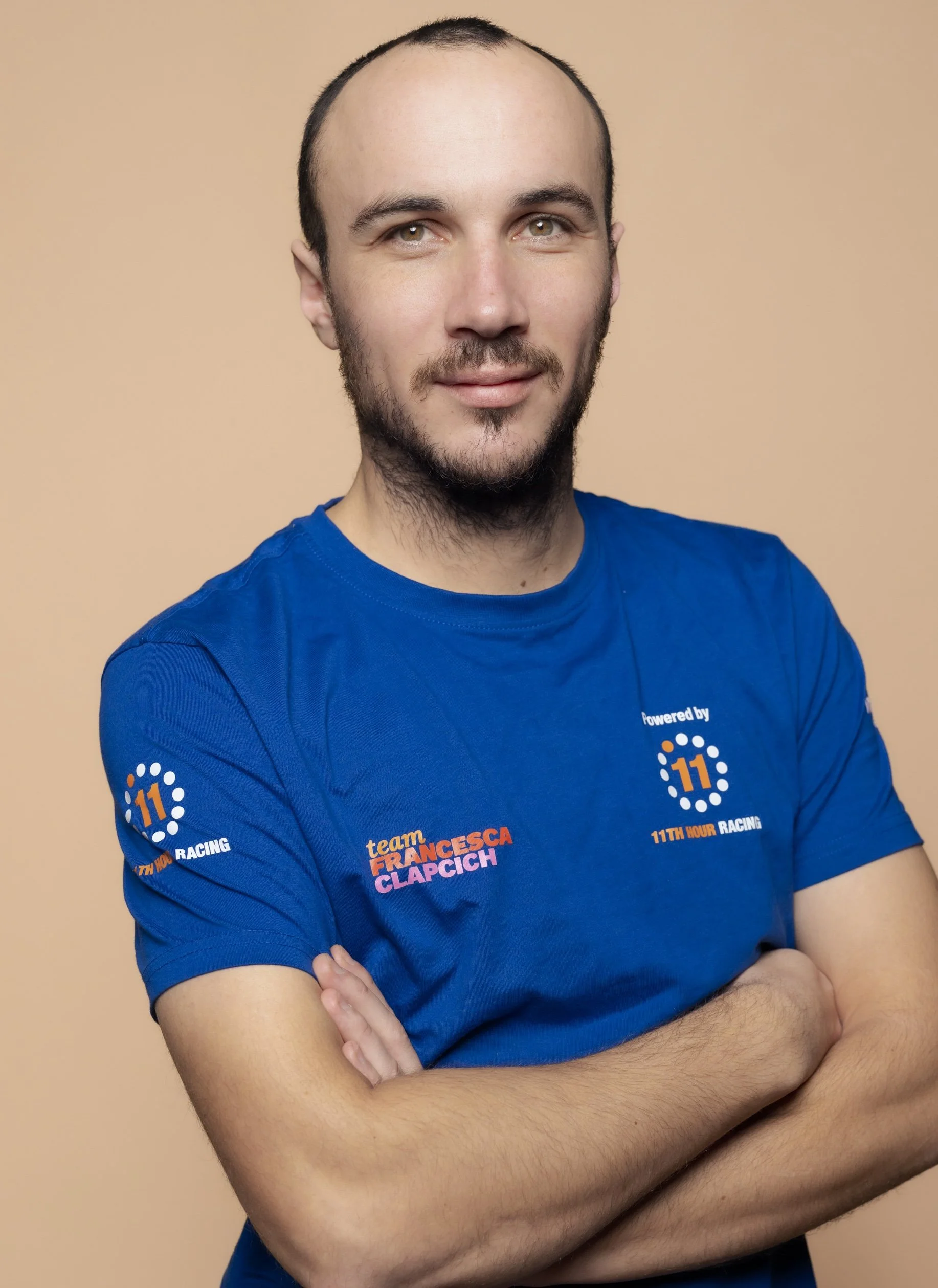 Portrait of a young man with a beard and short hair, wearing a blue team t-shirt with logos and text, standing with arms crossed against a beige background.