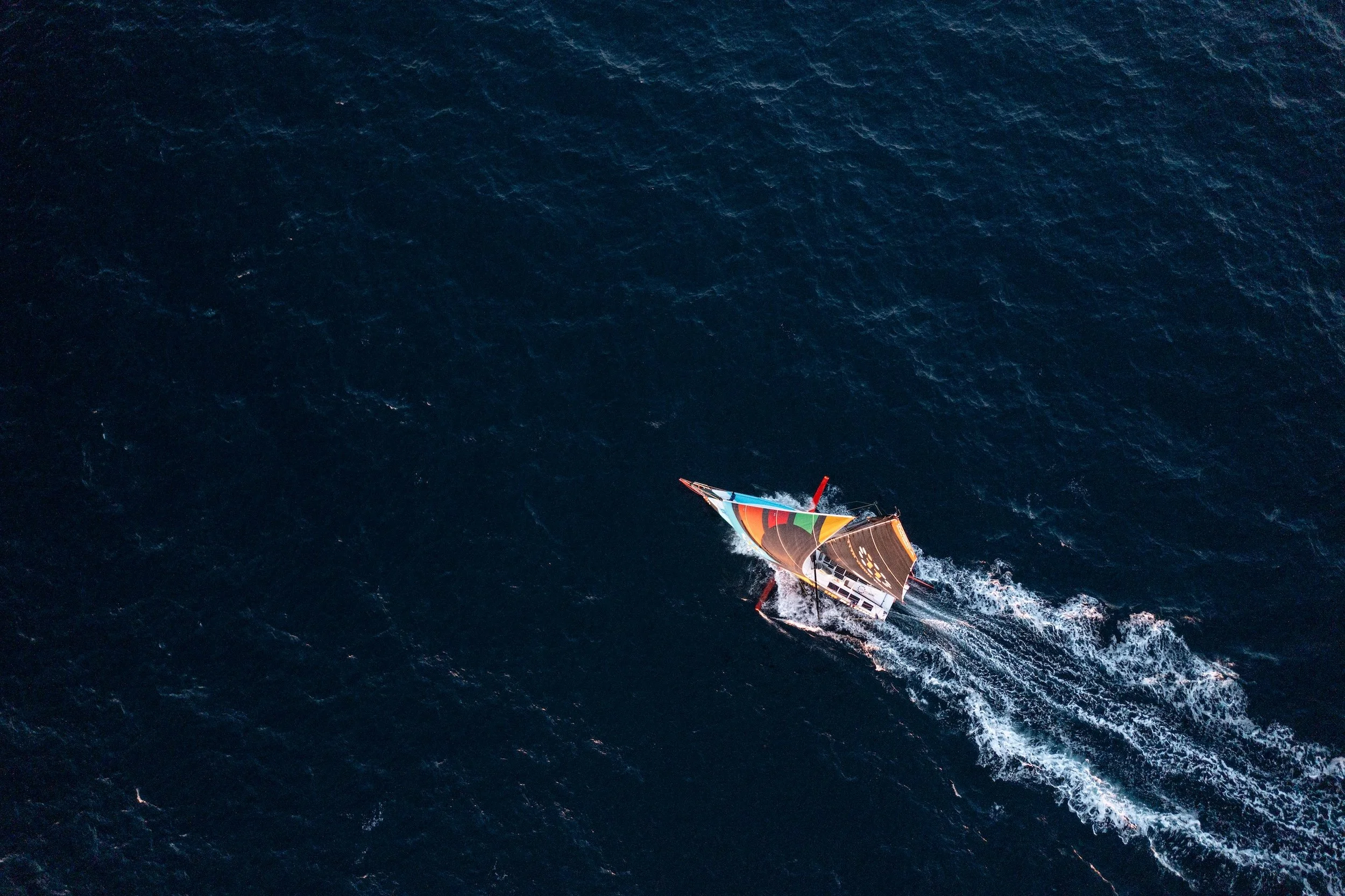 Overhead aerial view of a colorful sailing yacht navigating through dark blue ocean waters.