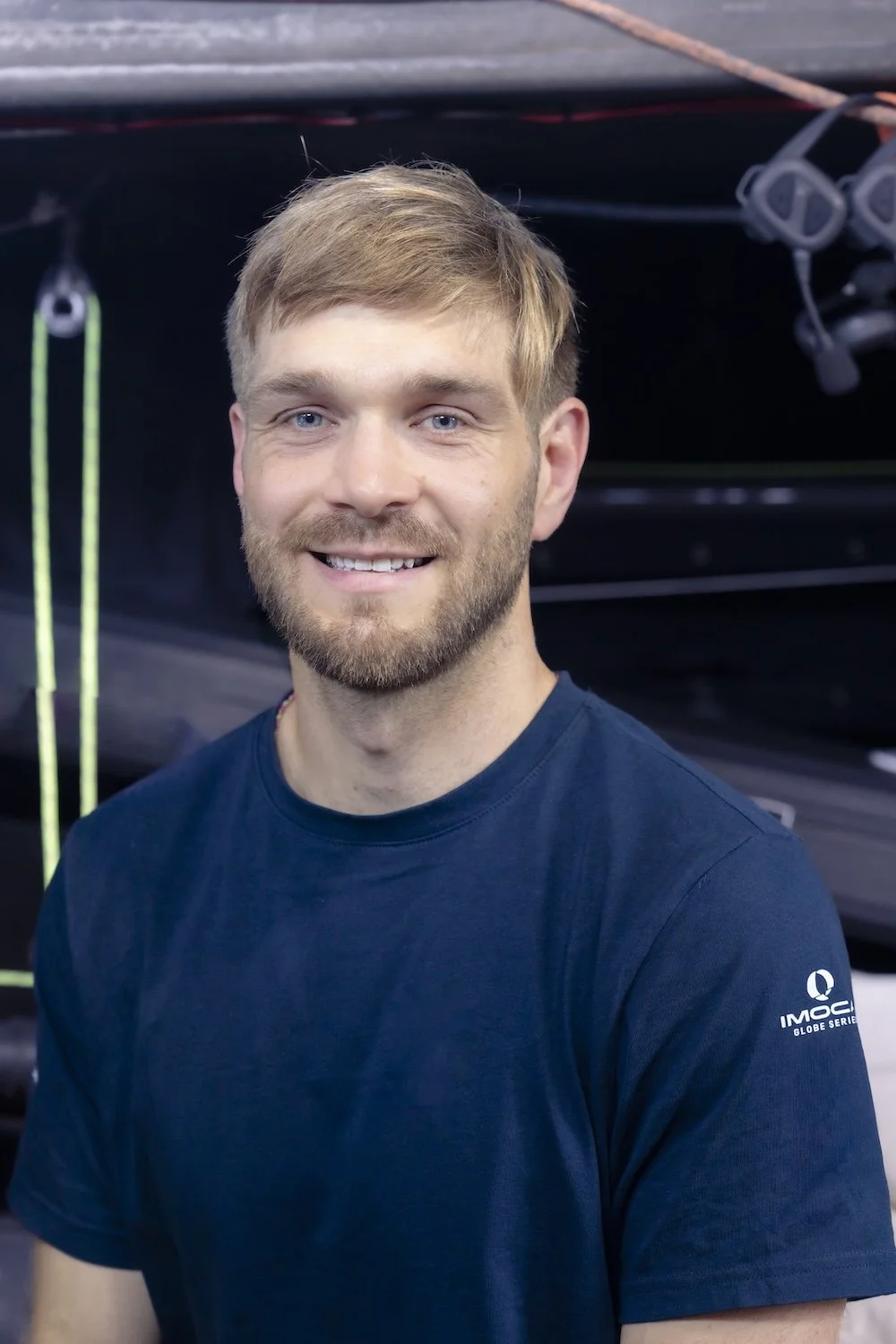 A smiling young man with short blond hair and a beard, wearing a navy blue Imoca logo t-shirt, standing in front of a boat with equipment.