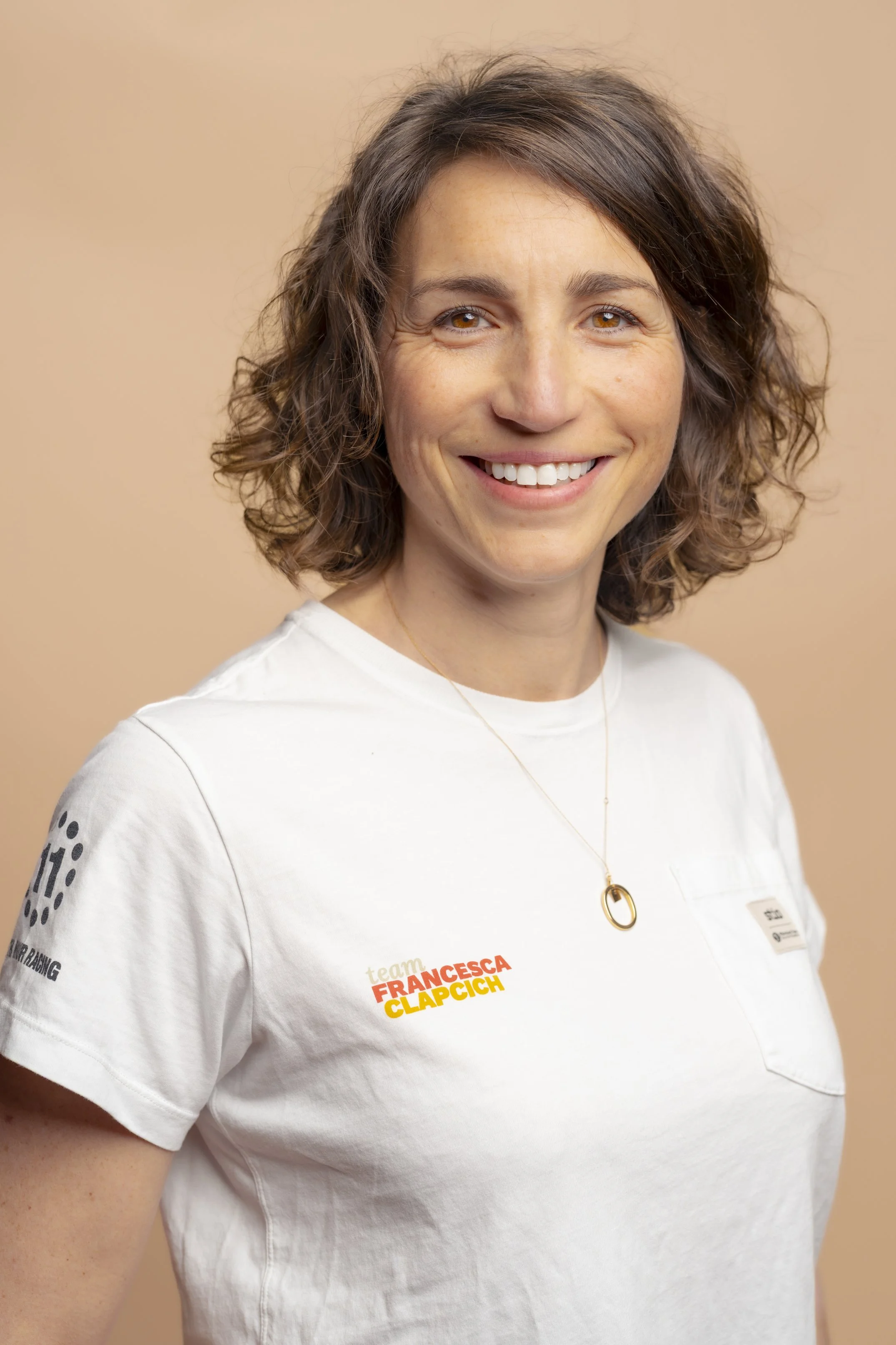 Portrait of a smiling woman with wavy brown hair, wearing a white T-shirt with the text 'team FRANCESCA CLAPCICH' and a gold necklace, against a beige background.