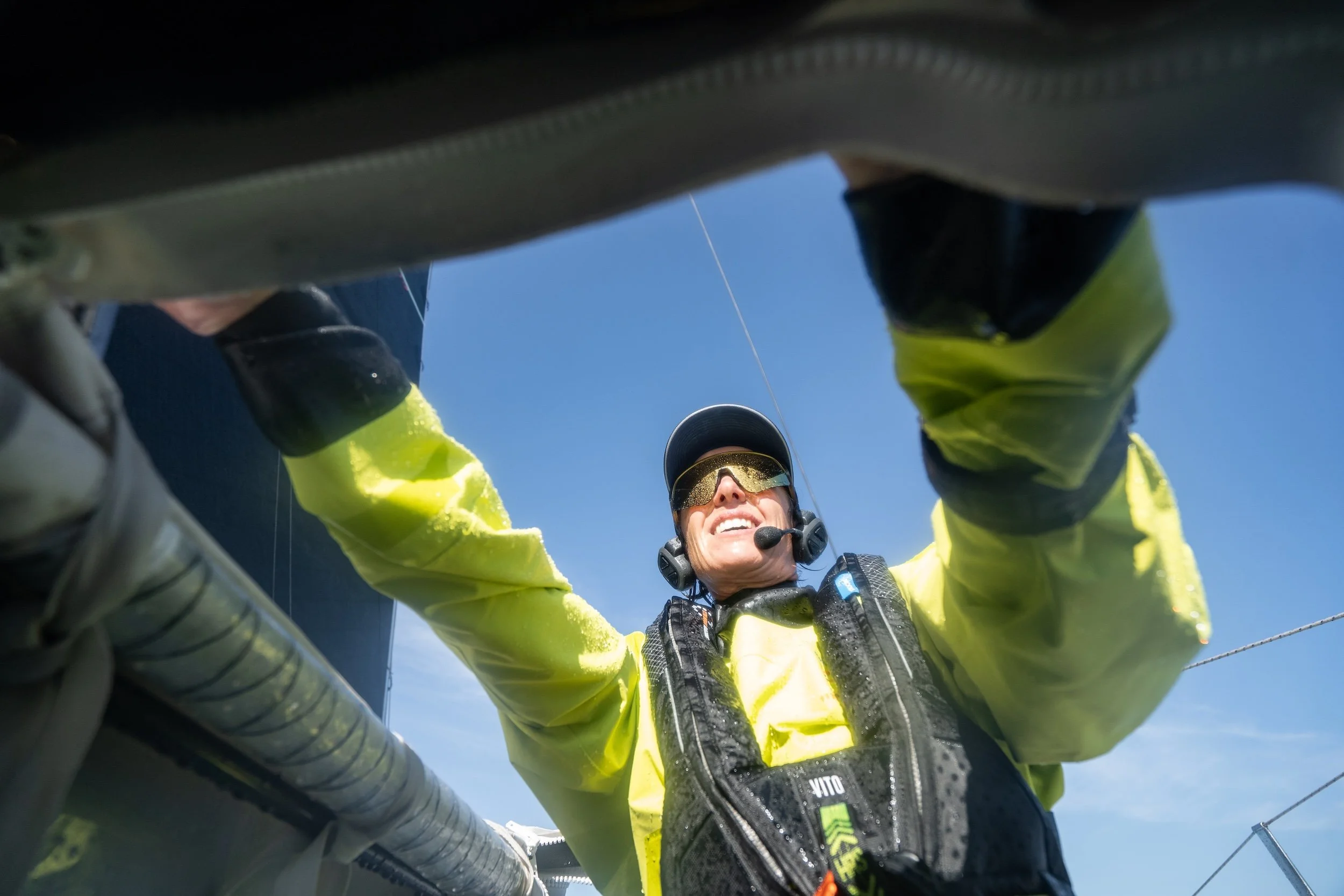 A person with a smile, wearing a yellow jacket, sunglasses, and a headset, is working on a boat or ship against a blue sky.