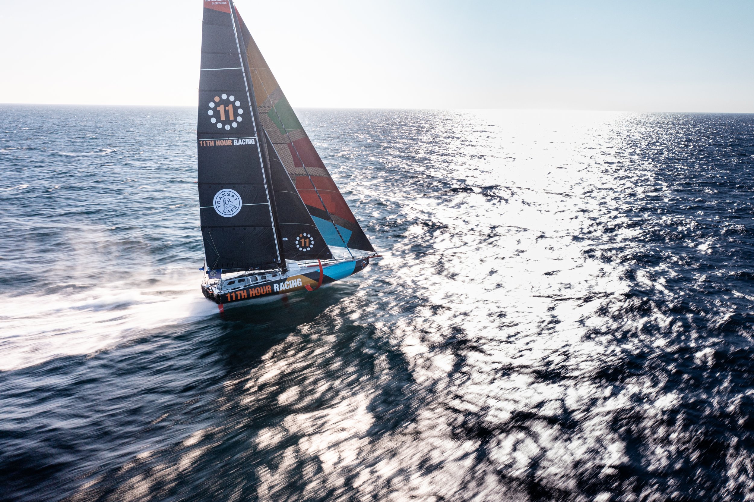 A racing sailboat with black, red, blue, and green sails on the open sea, creating a wake behind it during daylight.