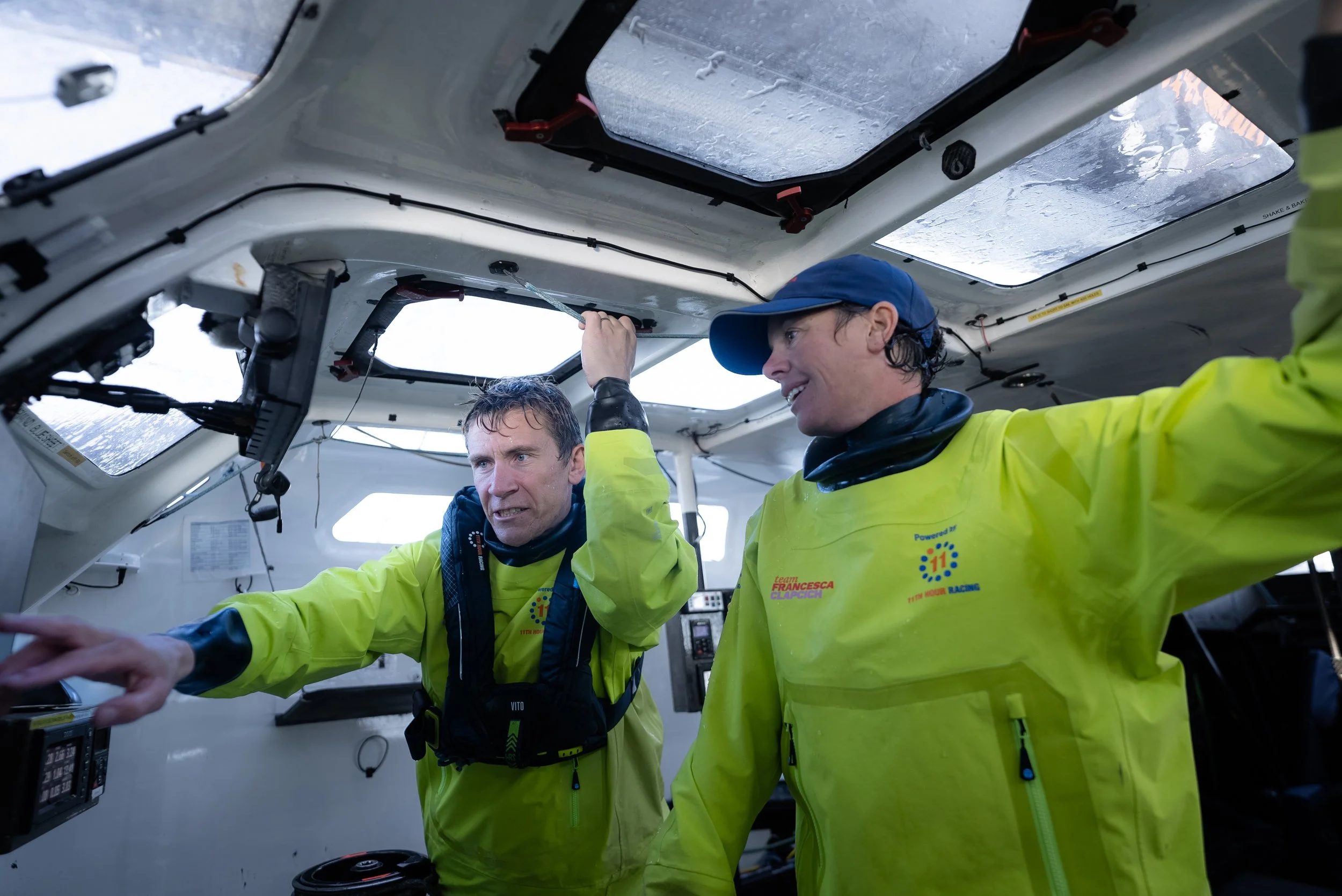 Two men in yellow racing suits inside a boat, inspecting equipment and speaking to each other, with windows covered in frost.