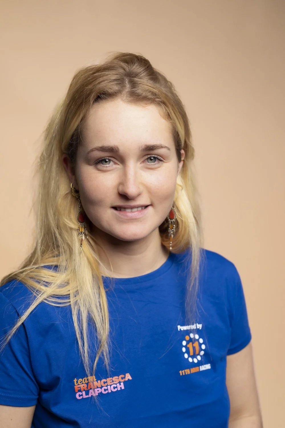 A young woman with blonde hair and blue eyes wearing a blue T-shirt with logos and text, standing against a beige background.
