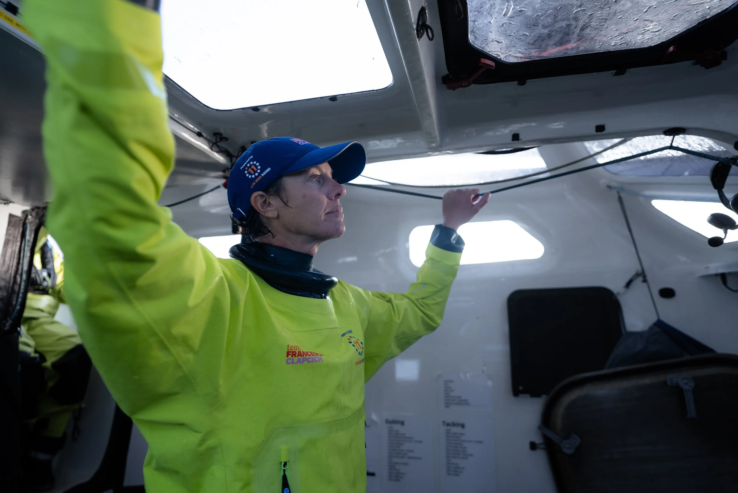 A woman wearing a yellow jacket and a blue cap inspecting or adjusting equipment inside a boat or yacht. She appears focused with her hand raised, touching wires or ropes near the ceiling of the vessel.