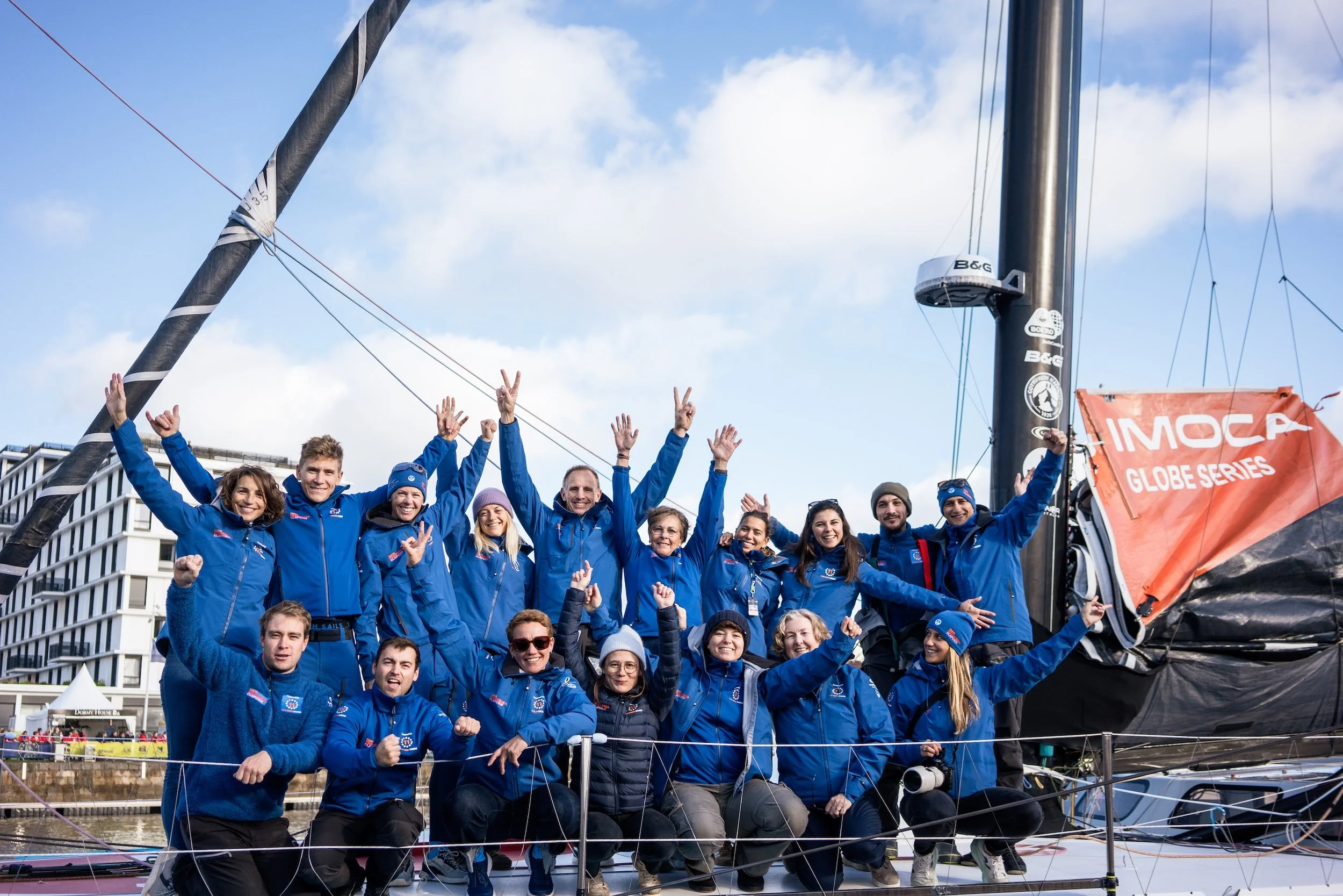 Team of sailors in blue jackets celebrating on a sailboat after winning a race, with a red banner reading 'IMoca Globe Series' and a modern building in the background.