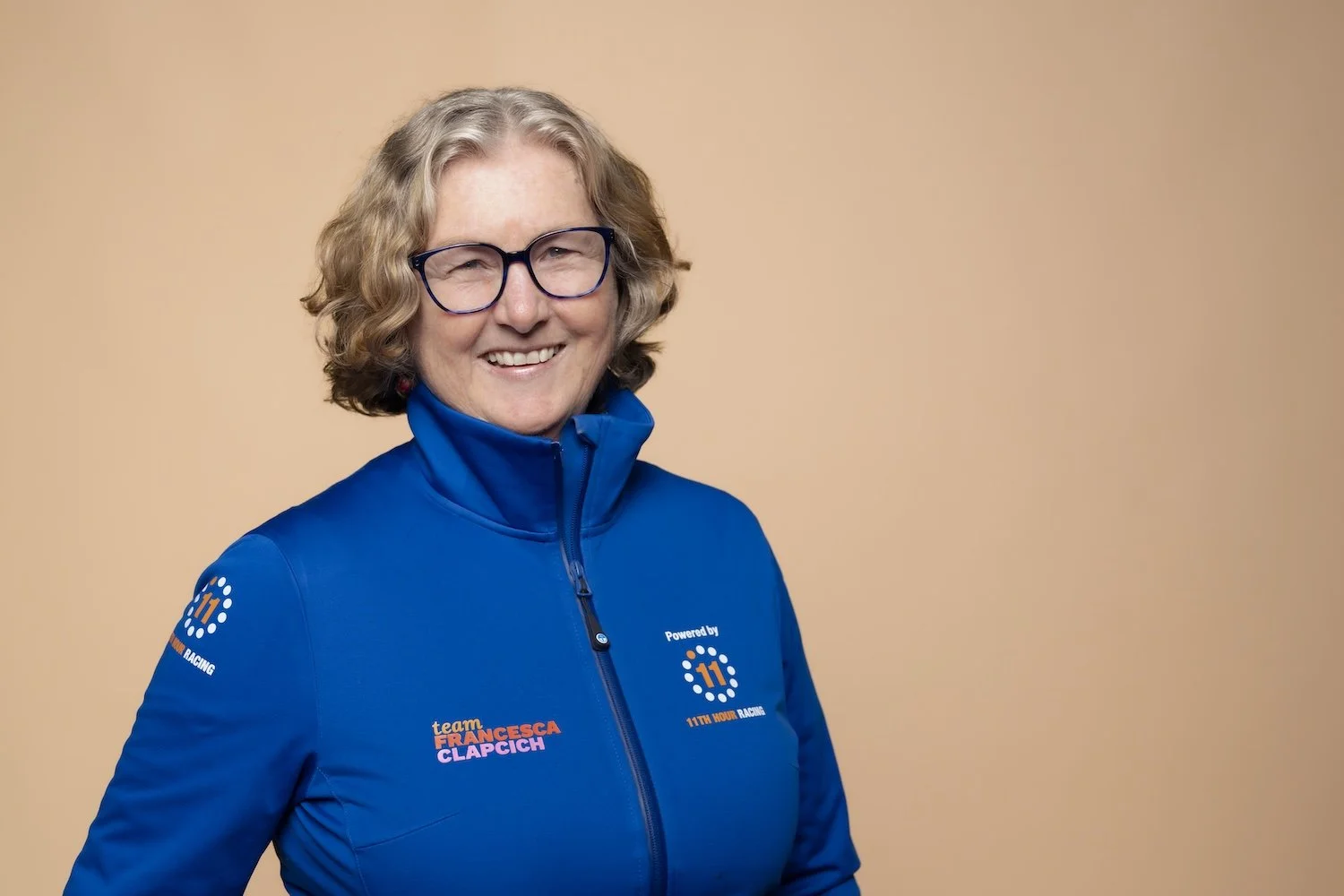 A smiling woman with curly gray hair and glasses wearing a blue jacket with logo and text.